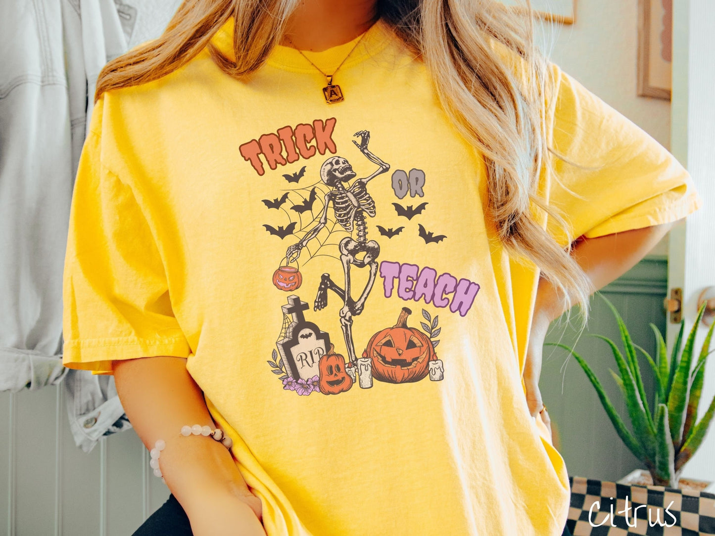 A woman wearing a cute, vintage citrus colored Comfort Colors T-shirt with text Trick or Teach in purple and orange font. There is a skeleton holding a pumpkin candy bucket dancing among spider webs and black bats over a tombstone and pumpkins.