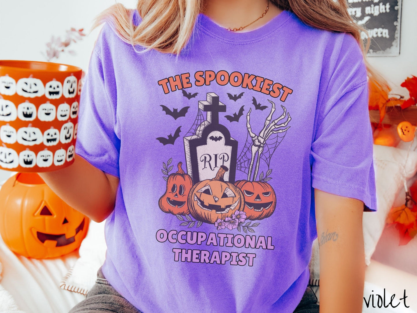 A woman wearing a cute, vintage violet colored Comfort Colors T-shirt with text The Spookiest Occupational Therapist. There are three carved pumpkins sitting in front of a tombstone with a skeleton arm coming out of the ground and bats in the air.