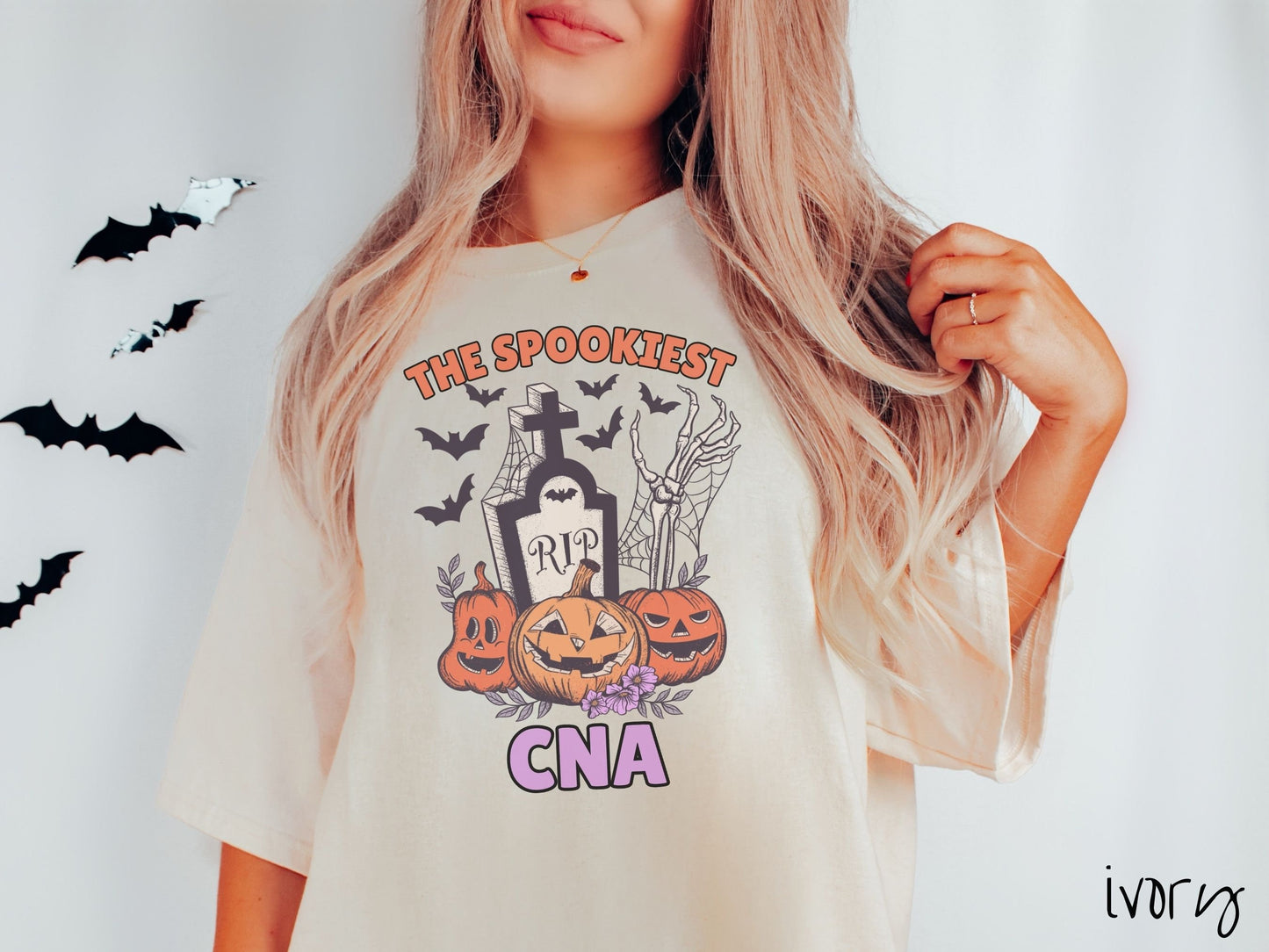 A woman wearing a cute, vintage ivory colored Comfort Colors T-shirt with text The Spookiest CNA. There are three carved pumpkins sitting in front of a tombstone with a skeleton arm coming out of the ground and bats in the air.