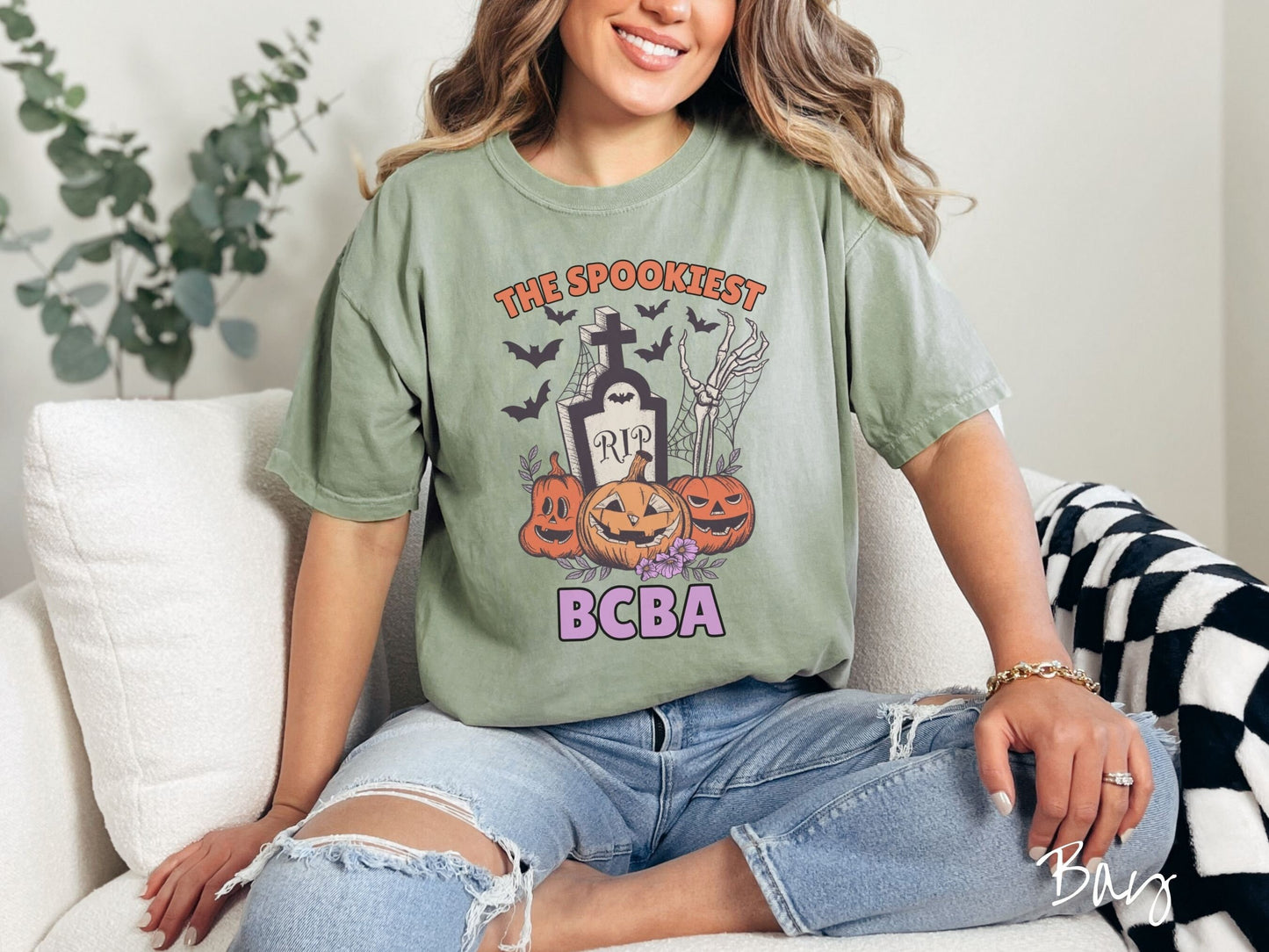 A woman wearing a cute, vintage bay colored Comfort Colors T-shirt with text The Spookiest BCBA. There are three pumpkins sitting in front of a tombstone with a skeleton arm coming out of the ground and bats in the air.
