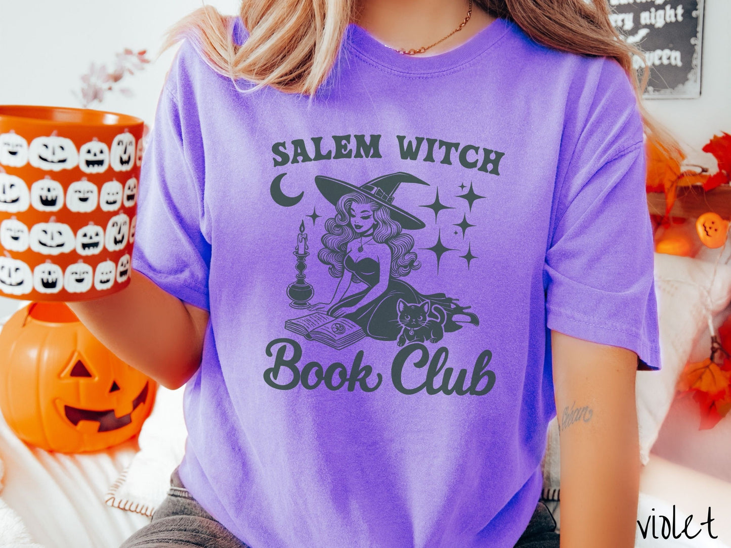 A woman wearing a cute, vintage violet colored Comfort Colors T-shirt with the text Salem Witch Book Club. In between the text is a young female witch reading a book on her knees surrounded by a candle, black cat, and stars and the moon.