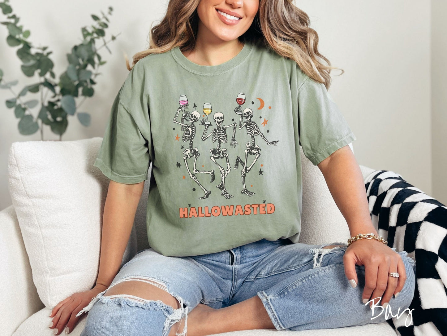 A woman wearing a cute, vintage bay colored Comfort Colors T-shirt with the text Hallowasted in orange font. Above are three female skeletons dancing while holding wine glasses with different colored wines and surrounded by stars.