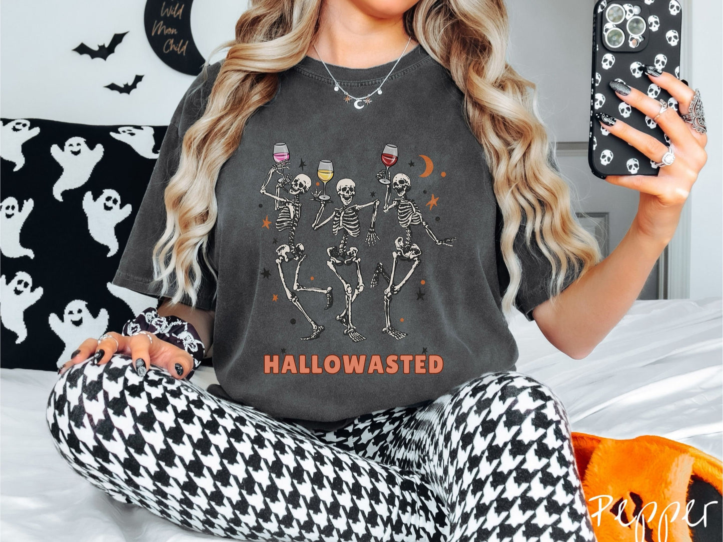 A woman wearing a cute, vintage pepper colored Comfort Colors T-shirt with the text Hallowasted in orange font. Above are three female skeletons dancing while holding wine glasses with different colored wines and surrounded by stars.