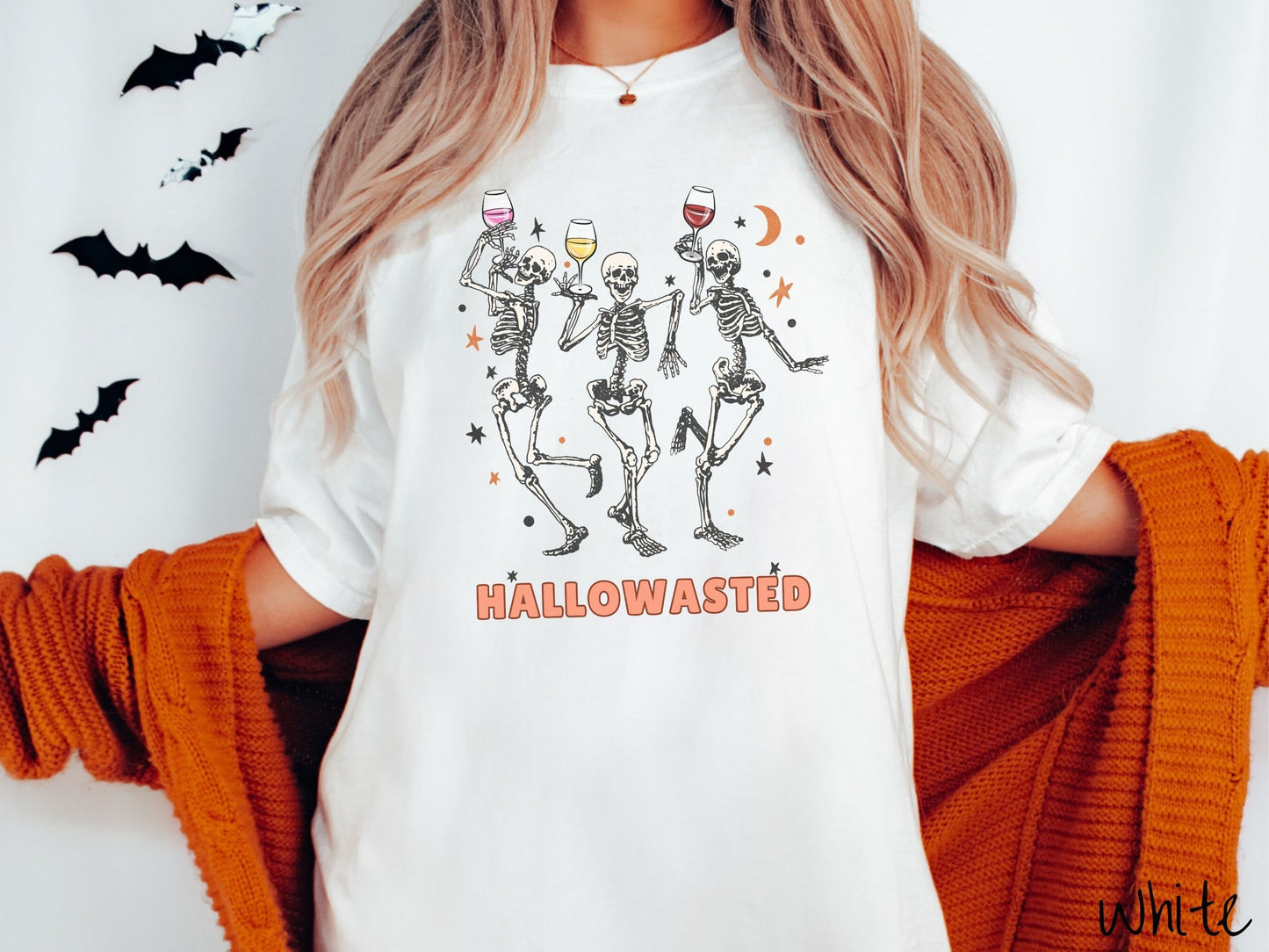 A woman wearing a cute, vintage white colored Comfort Colors T-shirt with the text Hallowasted in orange font. Above are three female skeletons dancing while holding wine glasses with different colored wines and surrounded by stars.