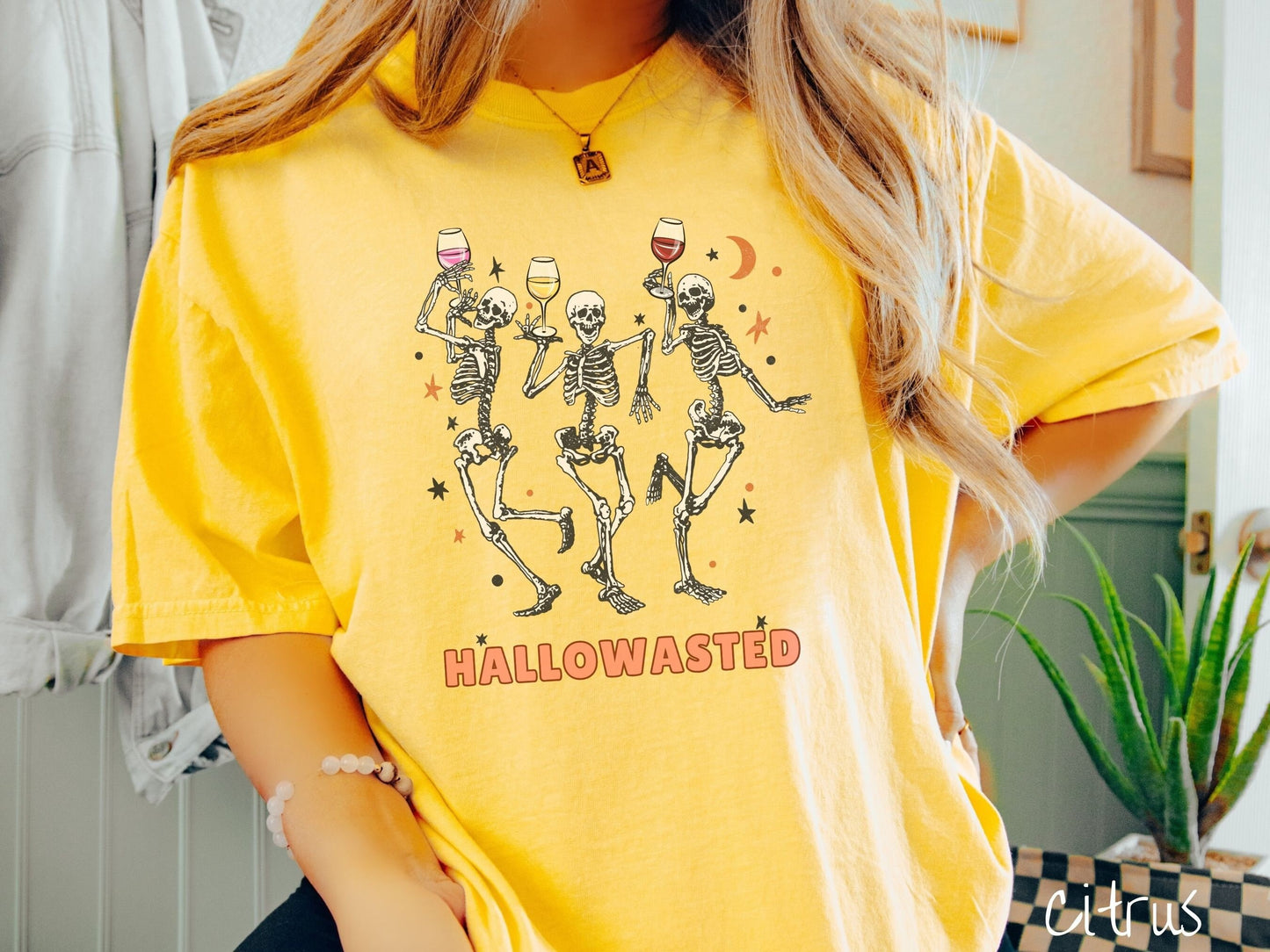 A woman wearing a cute, vintage citrus colored Comfort Colors T-shirt with the text Hallowasted in orange font. Above are three female skeletons dancing while holding wine glasses with different colored wines and surrounded by stars.
