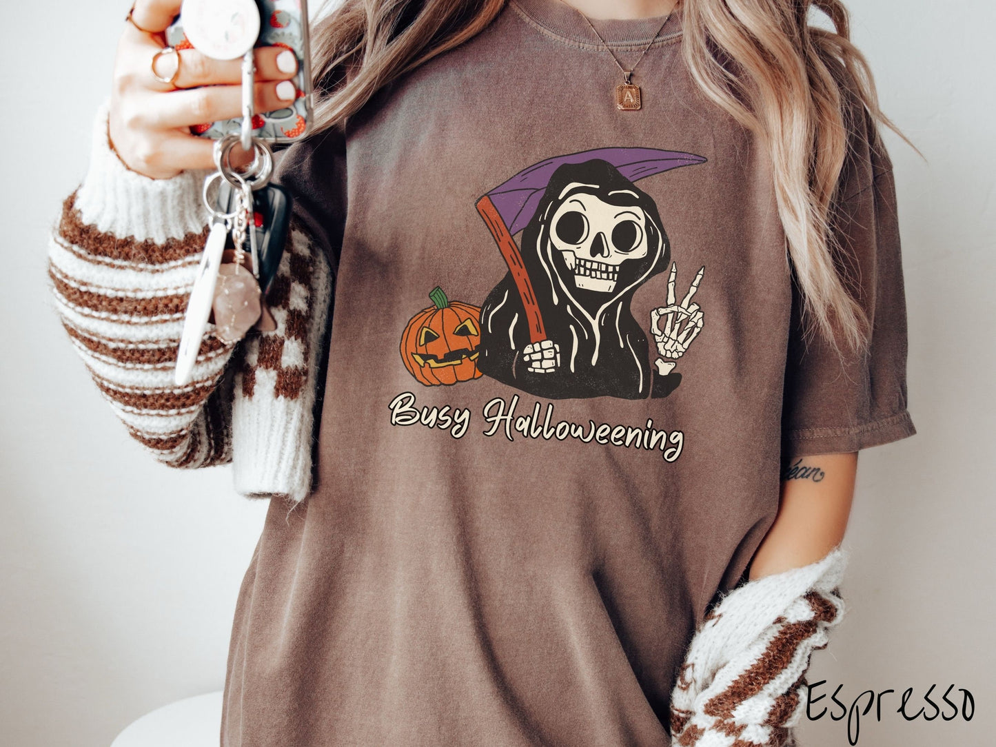 A woman wearing a cute, vintage espresso colored Comfort Colors T-shirt with text Busy Halloweening in white font. Above is a skeleton wearing a black hood holding up a peace sign in one hand and a scythe in the other with a carved pumpkin behind it.