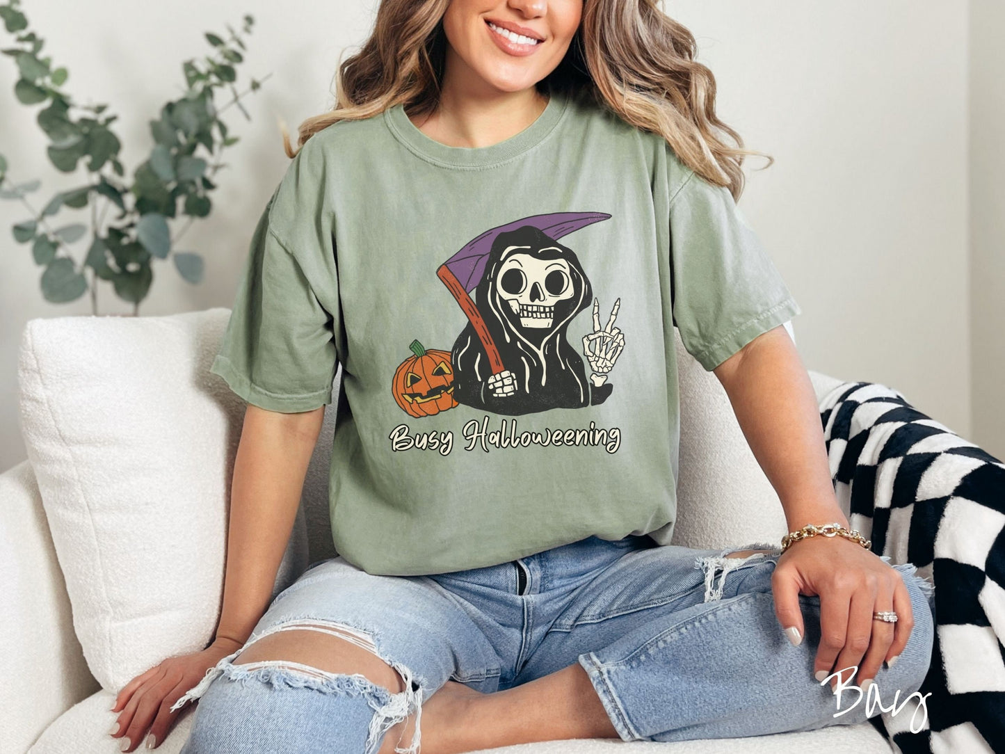 A woman wearing a cute, vintage bay colored Comfort Colors T-shirt with text Busy Halloweening in white font. Above is a skeleton wearing a black hood holding up a peace sign in one hand and a scythe in the other with a carved pumpkin behind it.