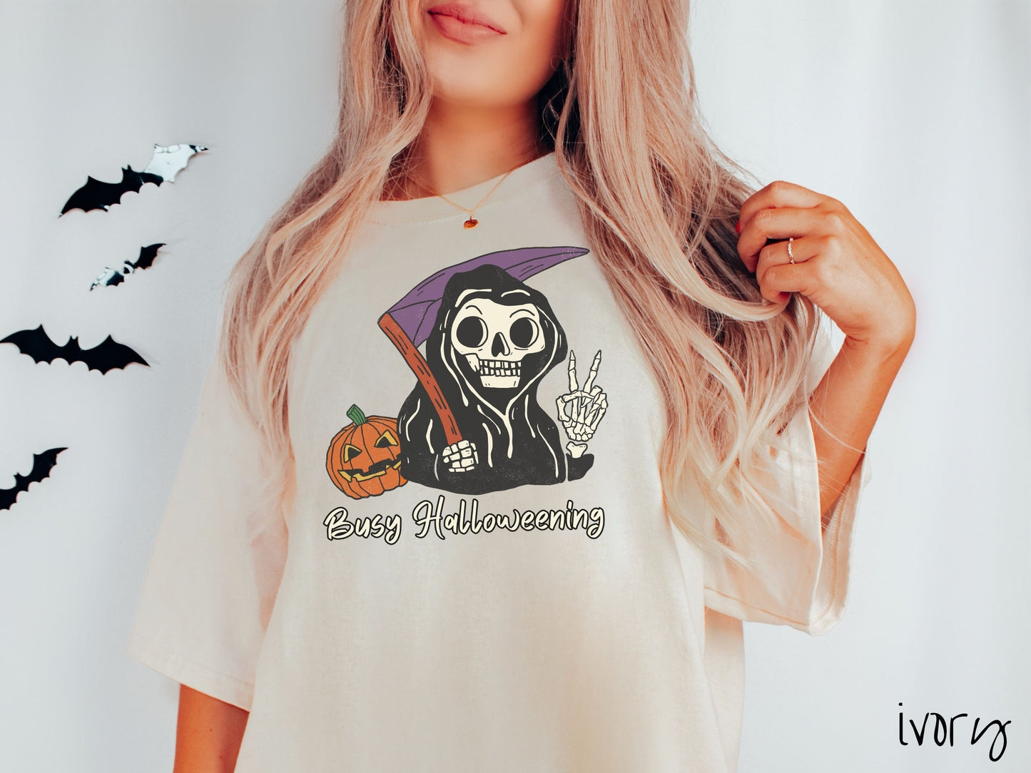 A woman wearing a cute, vintage ivory colored Comfort Colors T-shirt with text Busy Halloweening in white font. Above is a skeleton wearing a black hood holding up a peace sign in one hand and a scythe in the other with a carved pumpkin behind it.