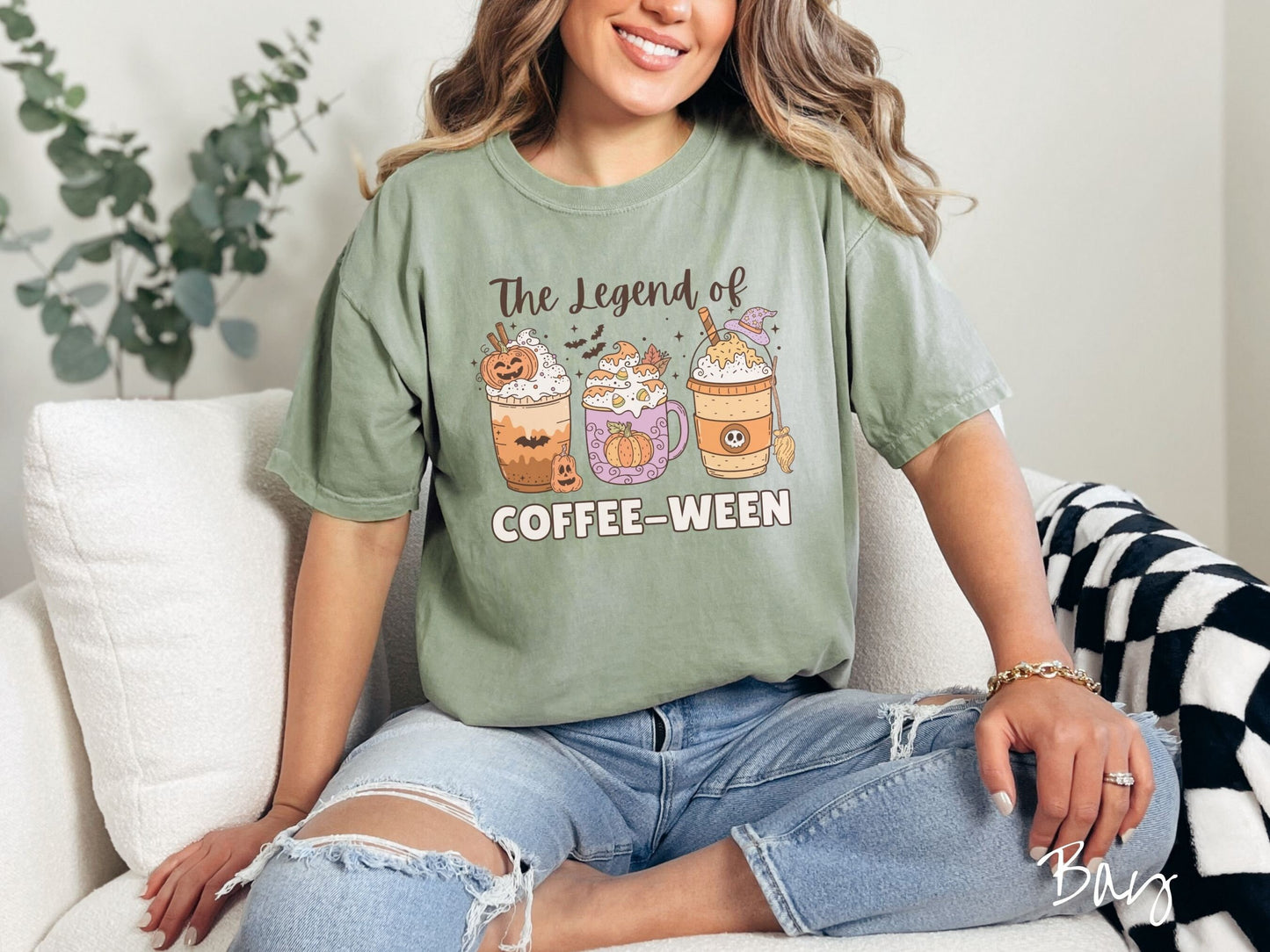 A woman wearing a cute, vintage bay colored Comfort Colors T-shirt with the text The legend of Coffee-ween. There are three iced coffees adorned with carved pumpkins, cinnamon sticks, whipped cream, bats, candy corn, whipped cream, and sprinkles.