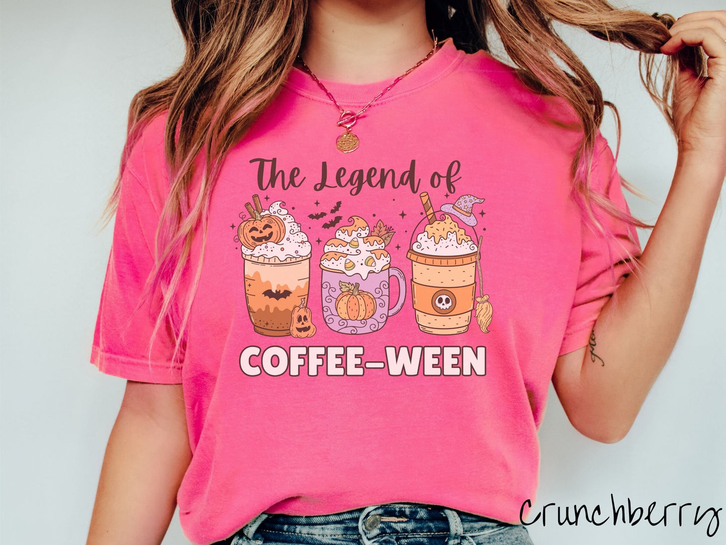 A woman wearing a cute, vintage crunchberry colored Comfort Colors T-shirt with text The legend of Coffee-ween. There are three iced coffees adorned with carved pumpkins, cinnamon sticks, whipped cream, bats, candy corn, whipped cream, and sprinkles.