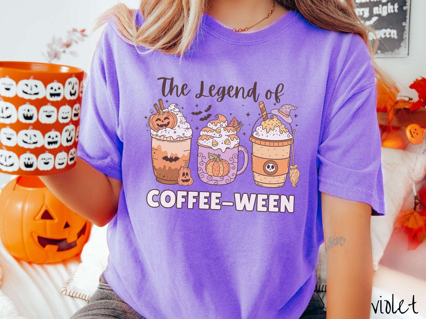 A woman wearing a cute, vintage violet colored Comfort Colors T-shirt with the text The legend of Coffee-ween. There are three iced coffees adorned with carved pumpkins, cinnamon sticks, whipped cream, bats, candy corn, whipped cream, and sprinkles.