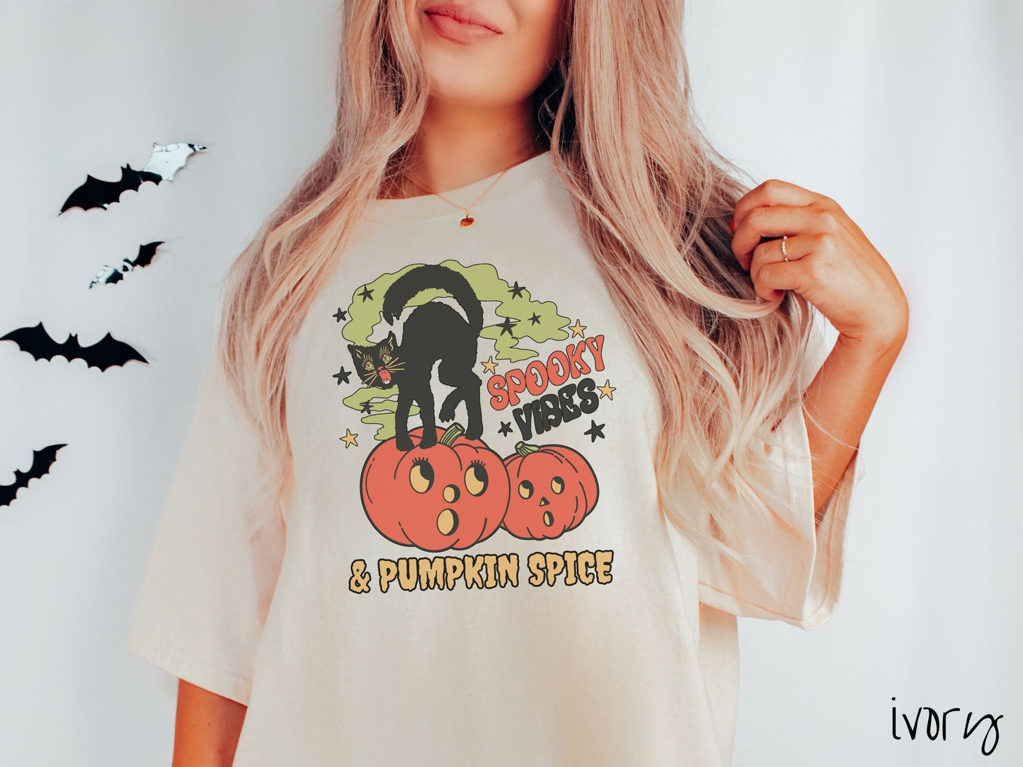 A woman wearing a cute, vintage ivory colored Comfort Colors T-shirt with the text Spooky Vibes & Pumpkin Spice in orange and black font. There is a black cat standing on an orange carved pumpkin and surrounded by green mist and twinkling stars.