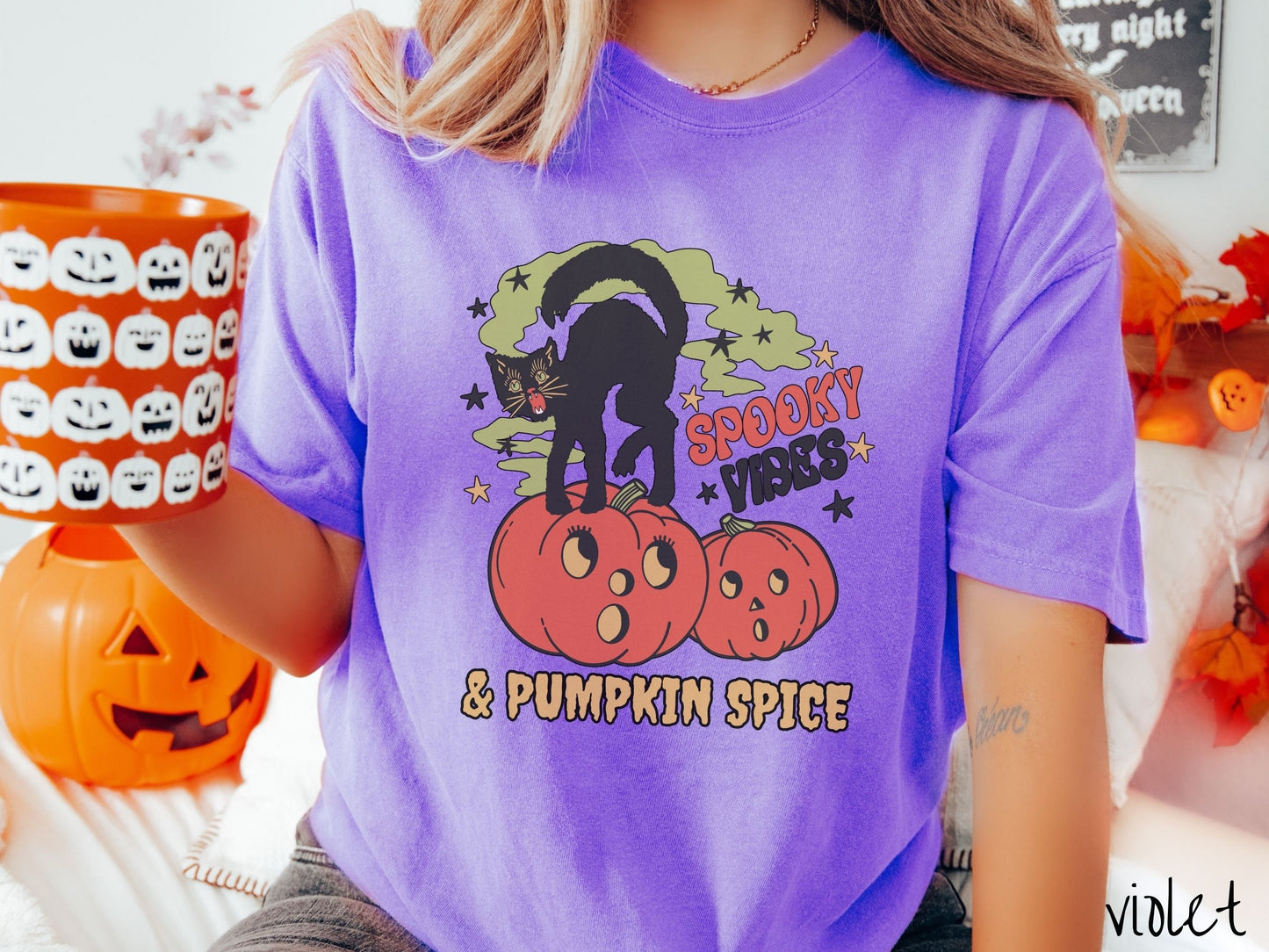 A woman wearing a cute, vintage violet colored Comfort Colors T-shirt with the text Spooky Vibes & Pumpkin Spice in orange and black font. There is a black cat standing on an orange carved pumpkin and surrounded by green mist and twinkling stars.