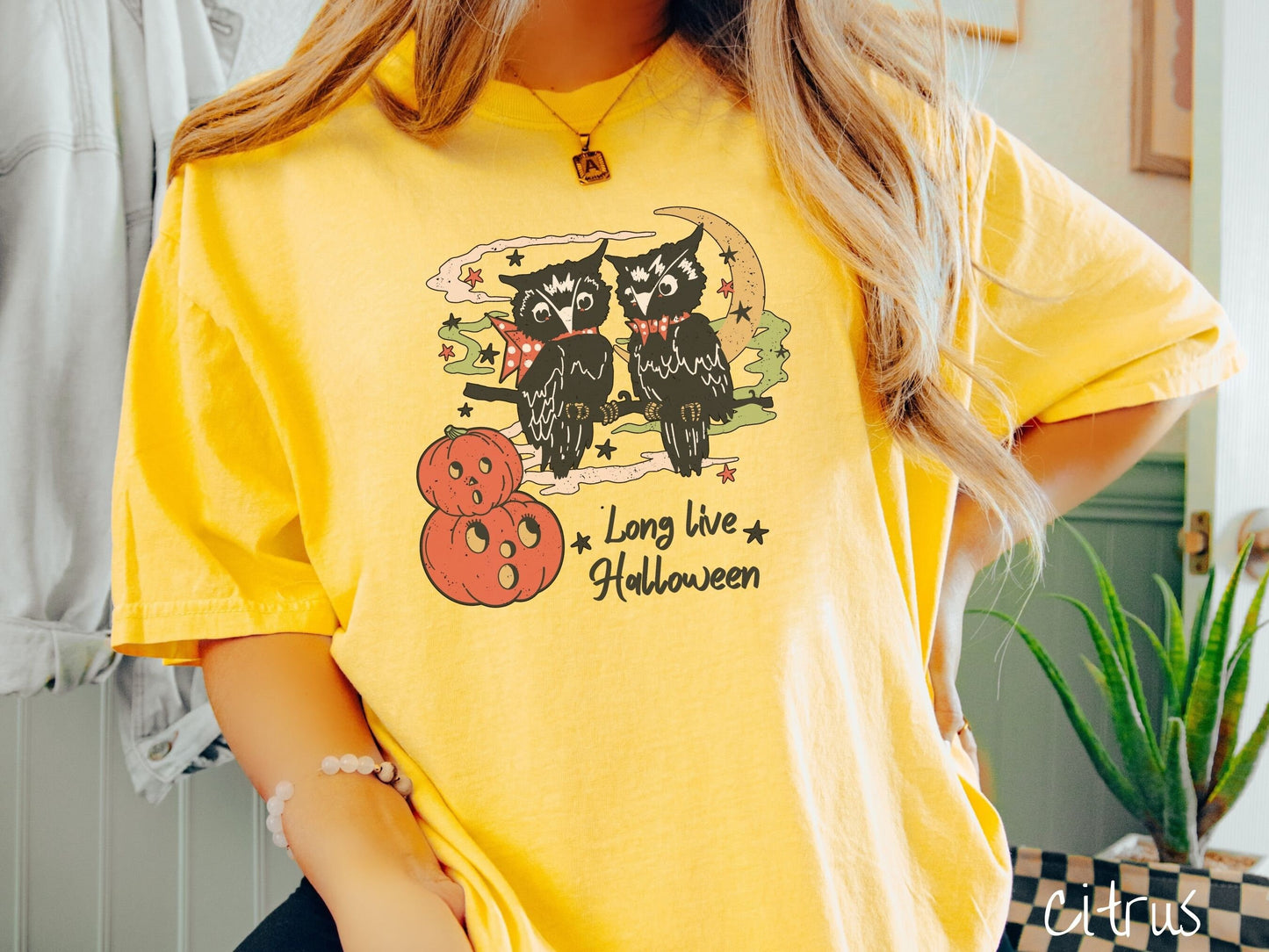 A woman wearing a cute, vintage citrus colored Comfort Colors T-shirt with the text Long Live Halloween in black font. Above are two black owls sitting on a tree branch by two scared pumpkins and surrounded by stars and the moon.