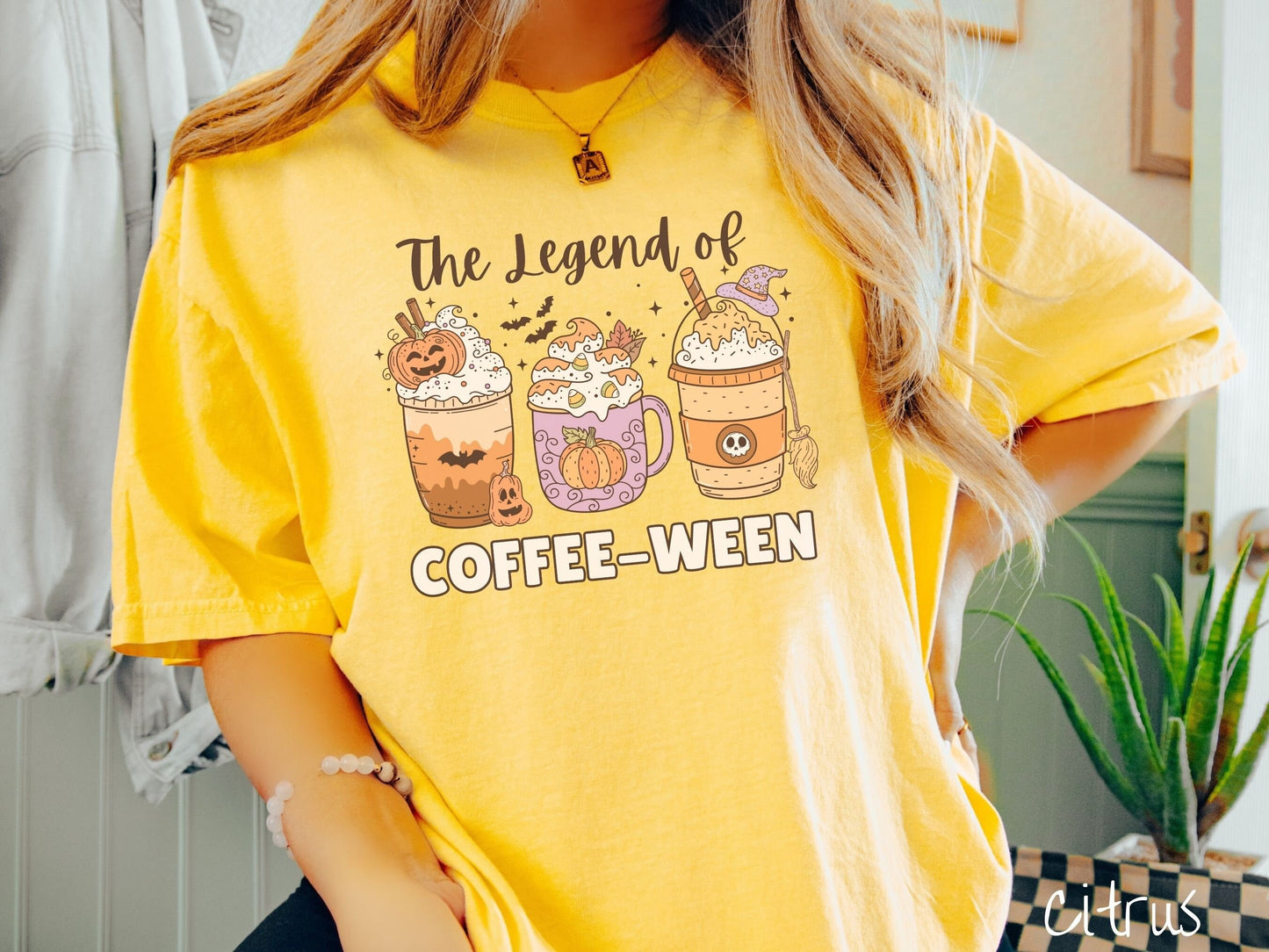 A woman wearing a cute, vintage citrus colored Comfort Colors T-shirt with the text The legend of Coffee-ween. There are three iced coffees adorned with carved pumpkins, cinnamon sticks, whipped cream, bats, candy corn, whipped cream, and sprinkles.
