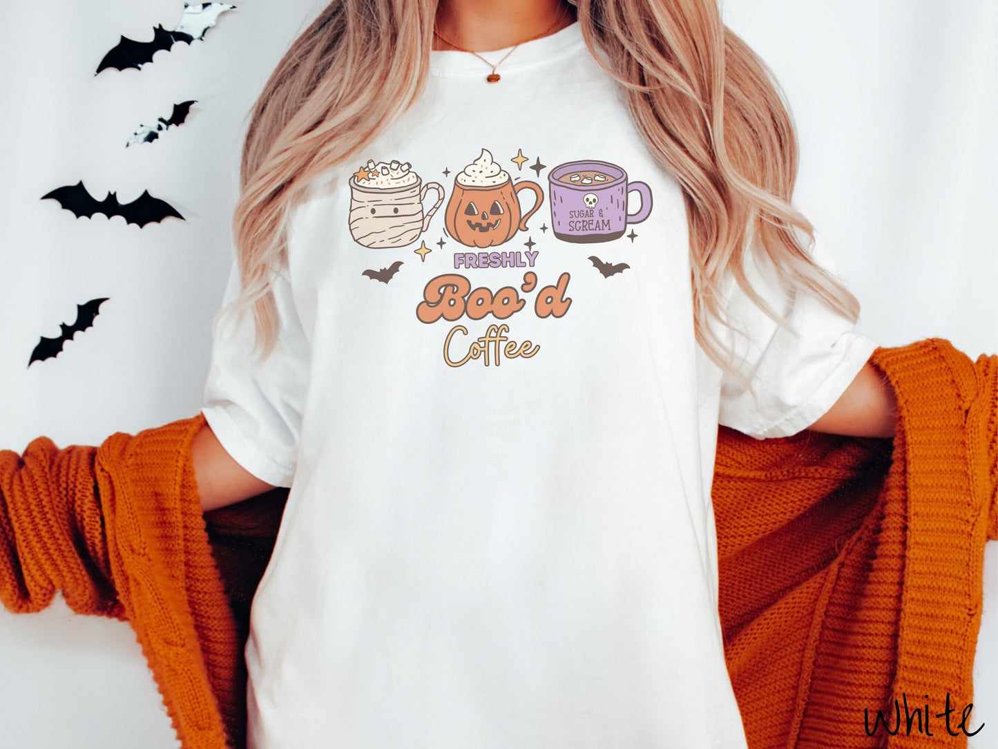 A woman wearing a cute, vintage white colored Comfort Colors T-shirt with the text Freshly Boo’d Coffee. Above are coffee mugs where one looks like a mummy with whipped cream, one a carved pumpkin mug, and the last is purple with a skull.