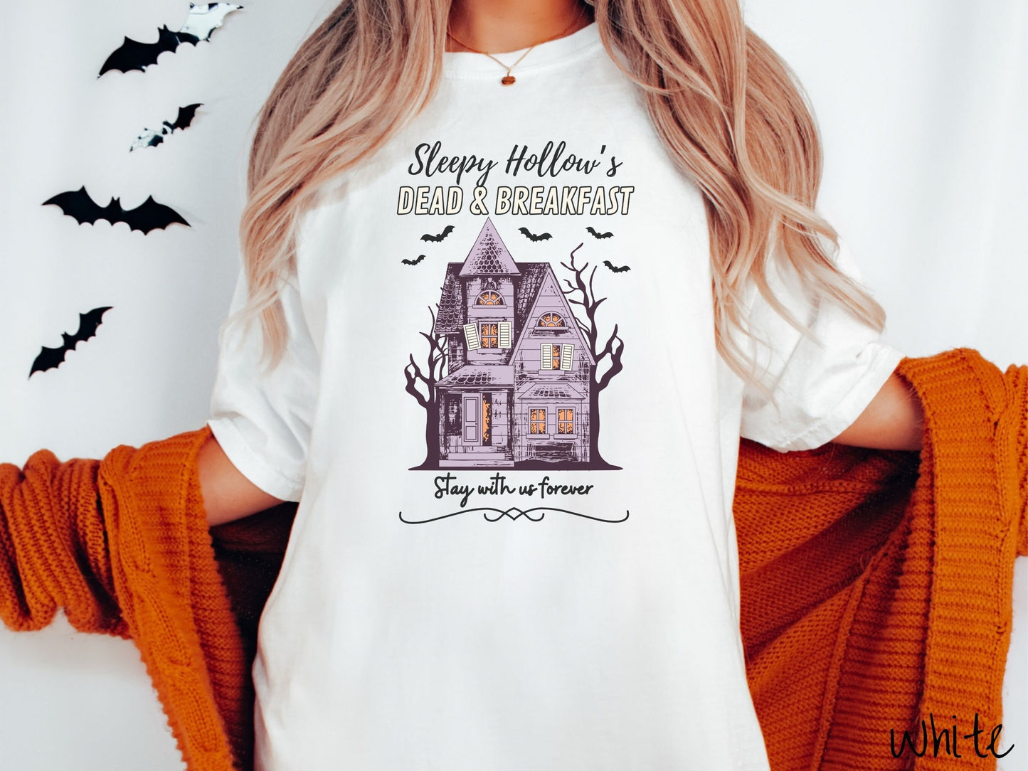 A woman wearing a cute, vintage white colored Comfort Colors T-shirt with the text Sleepy Hollow’s Dead & Breakfast above then Stay with us forever below. In between the text is a spooky haunted house surrounded by black bats.