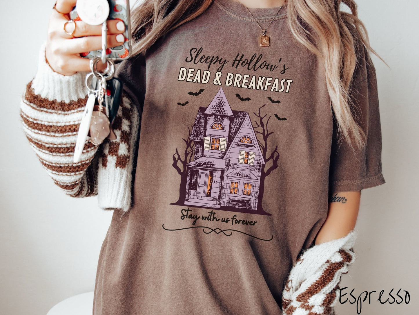 A woman wearing a cute, vintage espresso colored Comfort Colors T-shirt with the text Sleepy Hollow’s Dead & Breakfast above then Stay with us forever below. In between the text is a spooky haunted house surrounded by black bats.