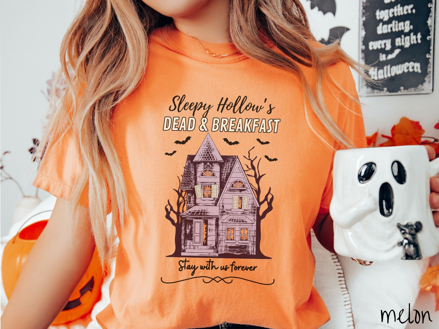 A woman wearing a cute, vintage melon colored Comfort Colors T-shirt with the text Sleepy Hollow’s Dead & Breakfast above then Stay with us forever below. In between the text is a spooky haunted house surrounded by black bats.