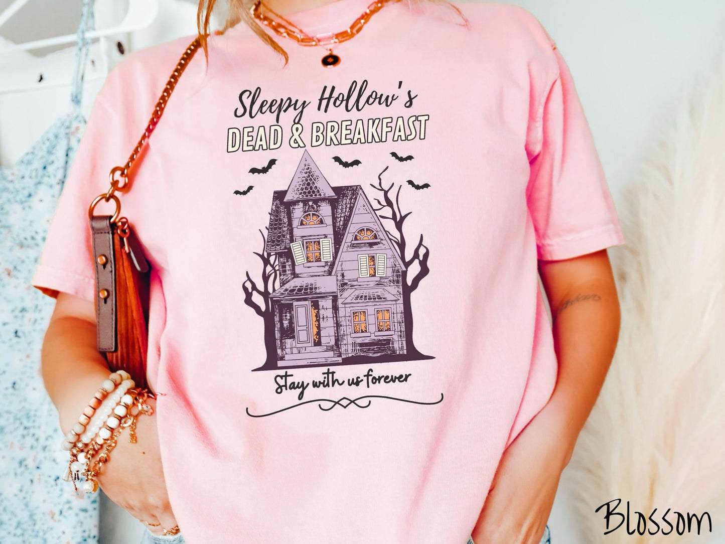 A woman wearing a cute, vintage blossom colored Comfort Colors T-shirt with the text Sleepy Hollow’s Dead & Breakfast above then Stay with us forever below. In between the text is a spooky haunted house surrounded by black bats.