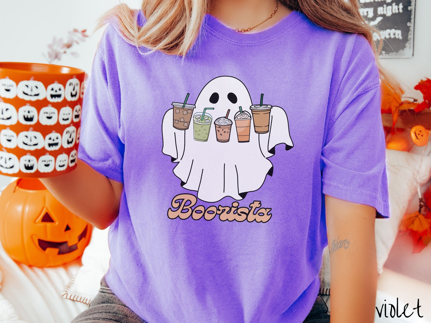 A woman wearing a cute, vintage violet colored Comfort Colors T-shirt with the text Boorista along the bottom. Above is a white ghost holding an iced coffee, a green matcha tea, a frappuccino, a pumpkin spiced latte, and an iced mocha latte.
