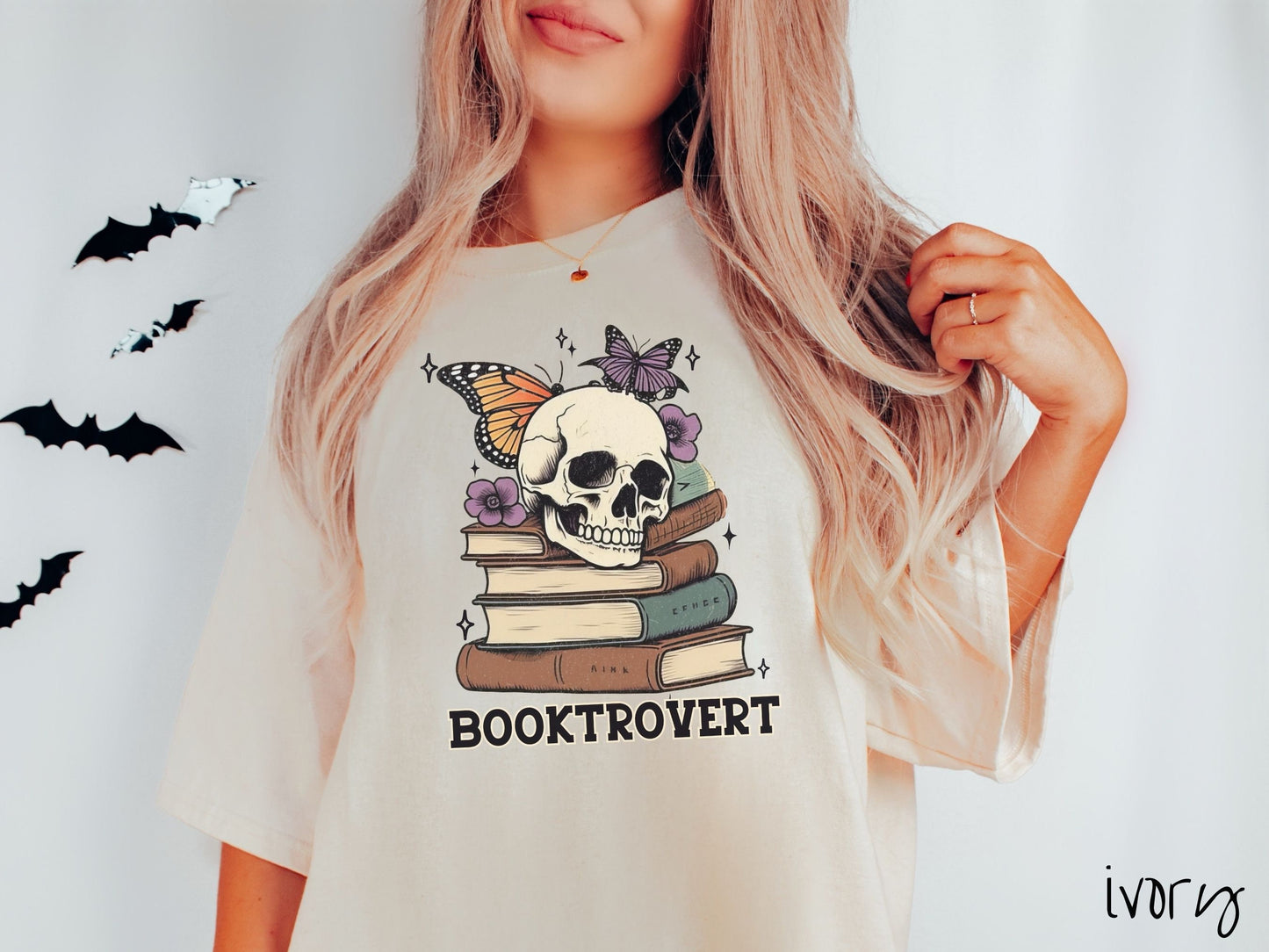 A woman wearing a cute, vintage ivory colored Comfort Colors T-shirt with the text Booktrovert in black font. Above are a stack of brown and green old books with a smiling skeleton skull sitting on top with colorful butterflies and flowers.