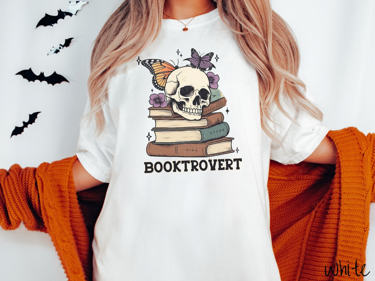 A woman wearing a cute, vintage white colored Comfort Colors T-shirt with the text Booktrovert in black font. Above are a stack of brown and green old books with a smiling skeleton skull sitting on top with colorful butterflies and flowers.