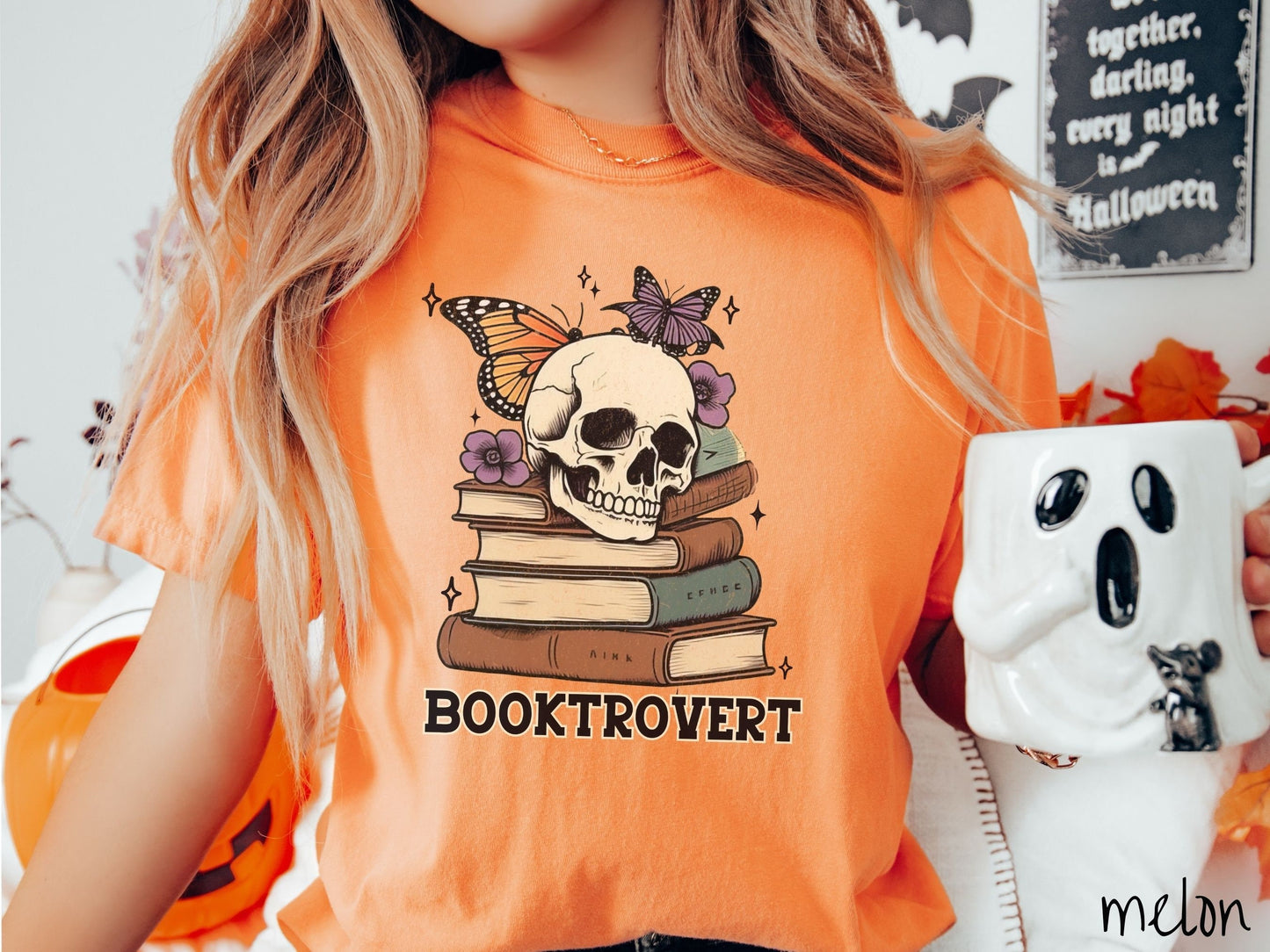 A woman wearing a cute, vintage melon colored Comfort Colors T-shirt with the text Booktrovert in black font. Above are a stack of brown and green old books with a smiling skeleton skull sitting on top with colorful butterflies and flowers.
