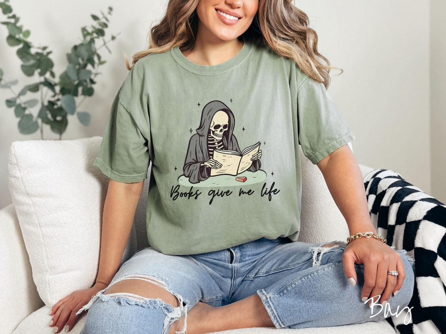 A woman wearing a cute, vintage bay colored Comfort Colors T-shirt with the text Books give me life in black cursive font. Above is a smiling skeleton wearing a black hood reading a book surrounded by twinkling stars.