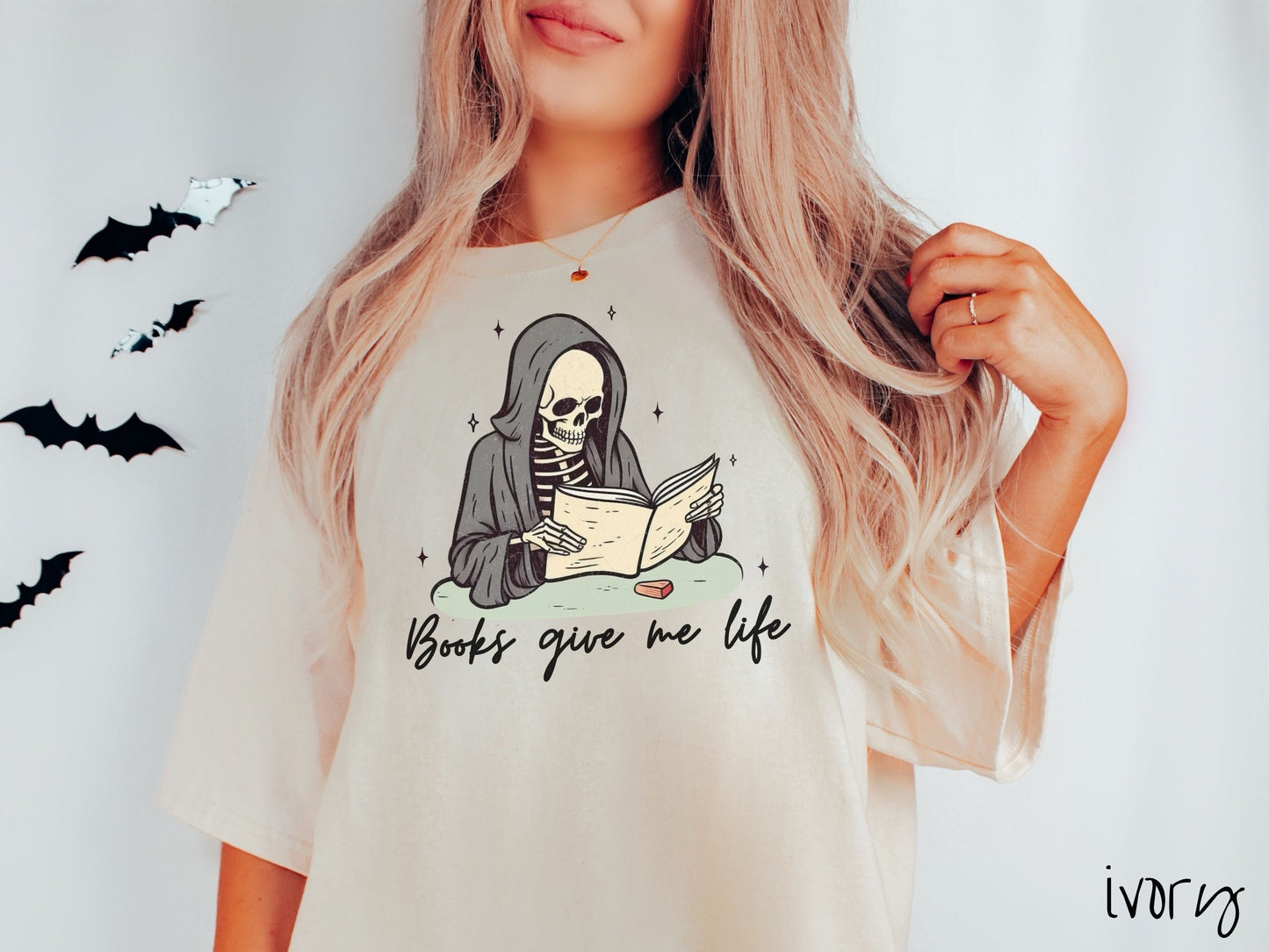 A woman wearing a cute, vintage ivory colored Comfort Colors T-shirt with the text Books give me life in black cursive font. Above is a smiling skeleton wearing a black hood reading a book surrounded by twinkling stars.
