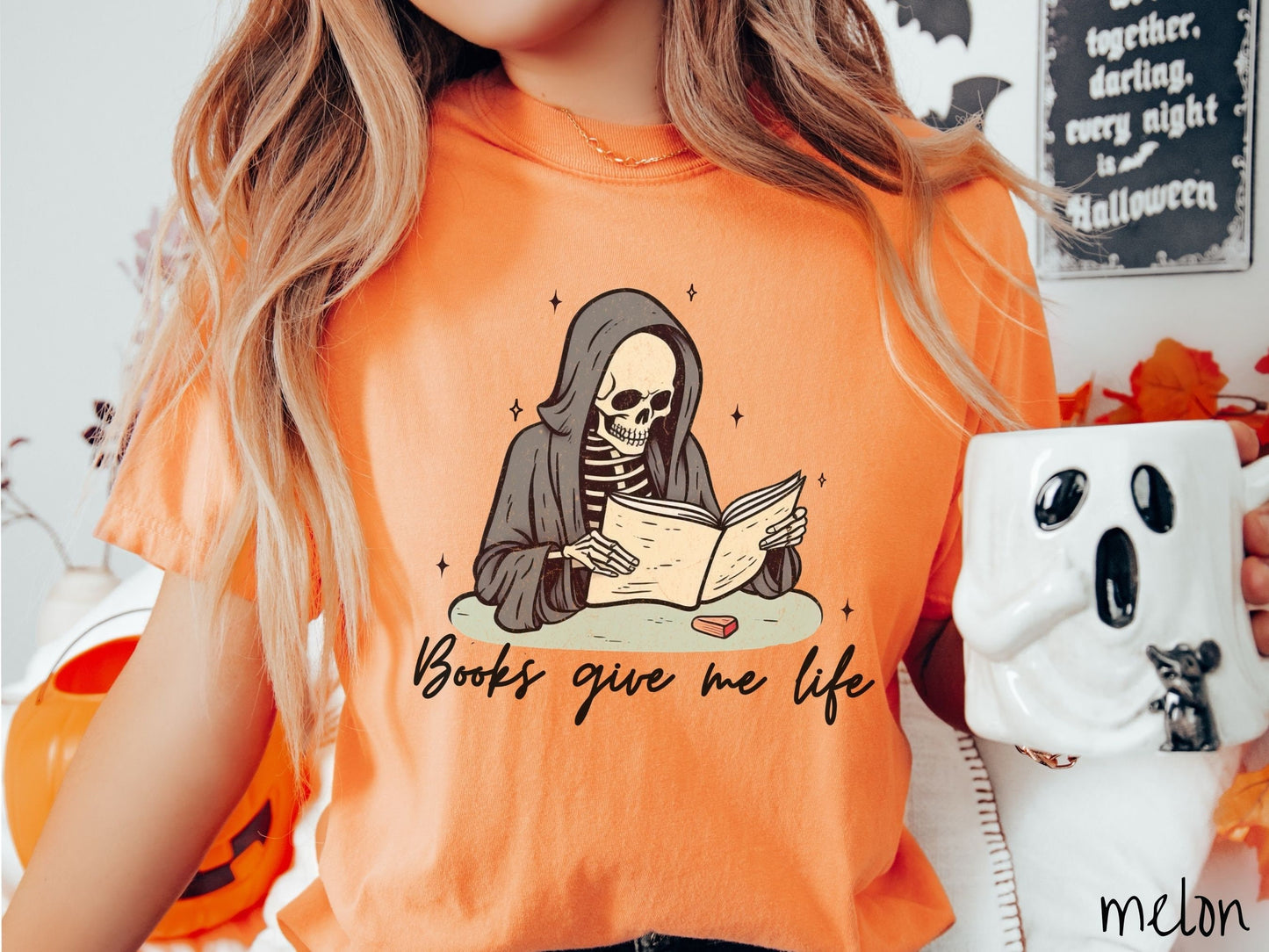 A woman wearing a cute, vintage melon colored Comfort Colors T-shirt with the text Books give me life in black cursive font. Above is a smiling skeleton wearing a black hood reading a book surrounded by twinkling stars.