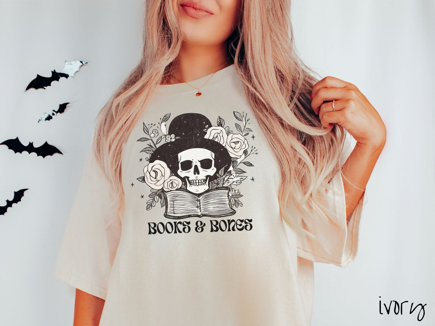A woman wearing a cute, vintage ivory colored Comfort Colors T-shirt with the text Books & Bones in black font. Above is a smiling skeleton wearing a black top hat surrounded by roses and leaves, and hovering above a thick, leather bound book.