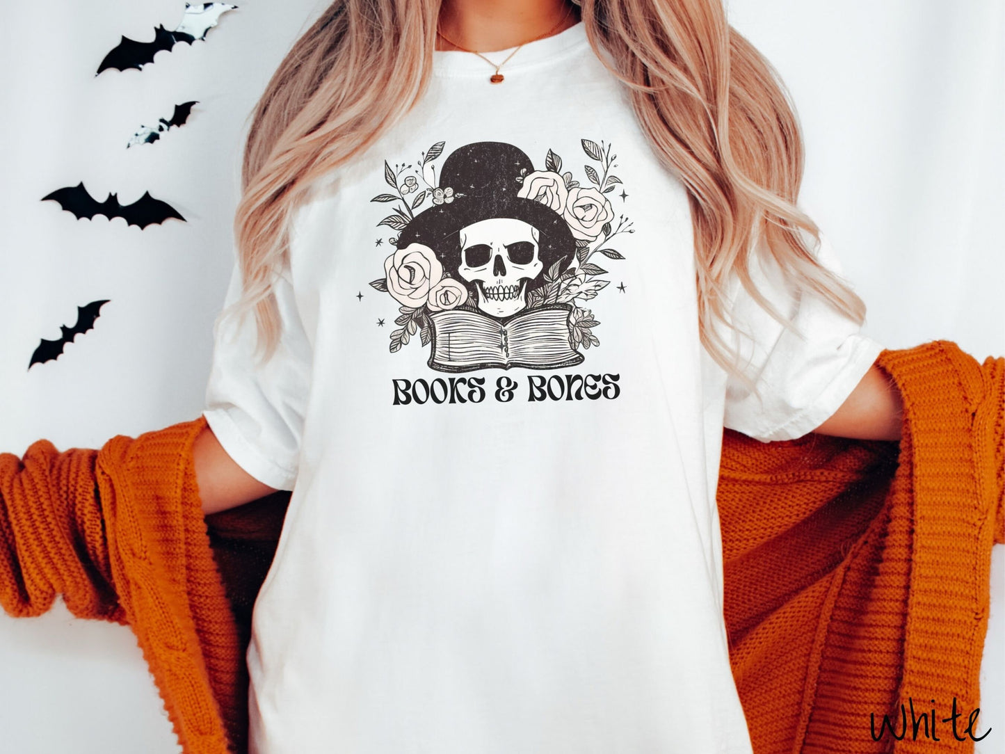 A woman wearing a cute, vintage white colored Comfort Colors T-shirt with the text Books & Bones in black font. Above is a smiling skeleton wearing a black top hat surrounded by roses and leaves, and hovering above a thick, leather bound book.