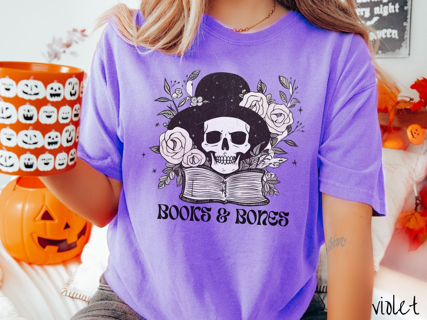A woman wearing a cute, vintage violet colored Comfort Colors T-shirt with the text Books & Bones in black font. Above is a smiling skeleton wearing a black top hat surrounded by roses and leaves, and hovering above a thick, leather bound book.
