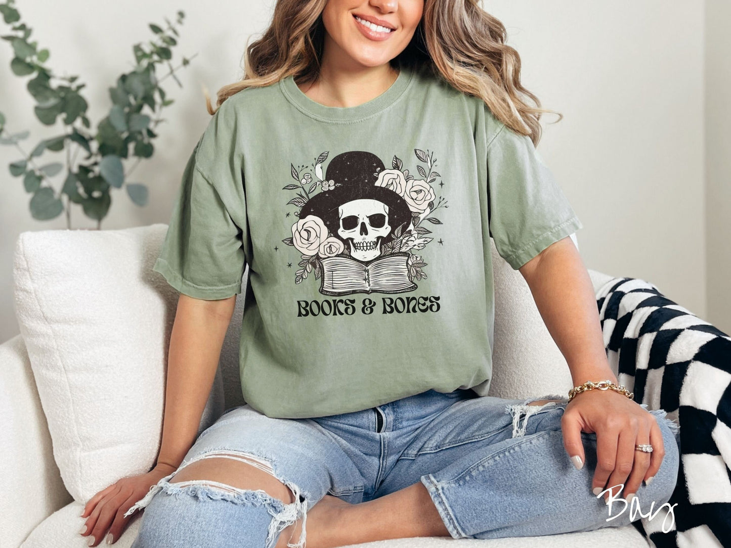A woman wearing a cute, vintage bay colored Comfort Colors T-shirt with the text Books & Bones in black font. Above is a smiling skeleton wearing a black top hat surrounded by roses and leaves, and hovering above a thick, leather bound book.