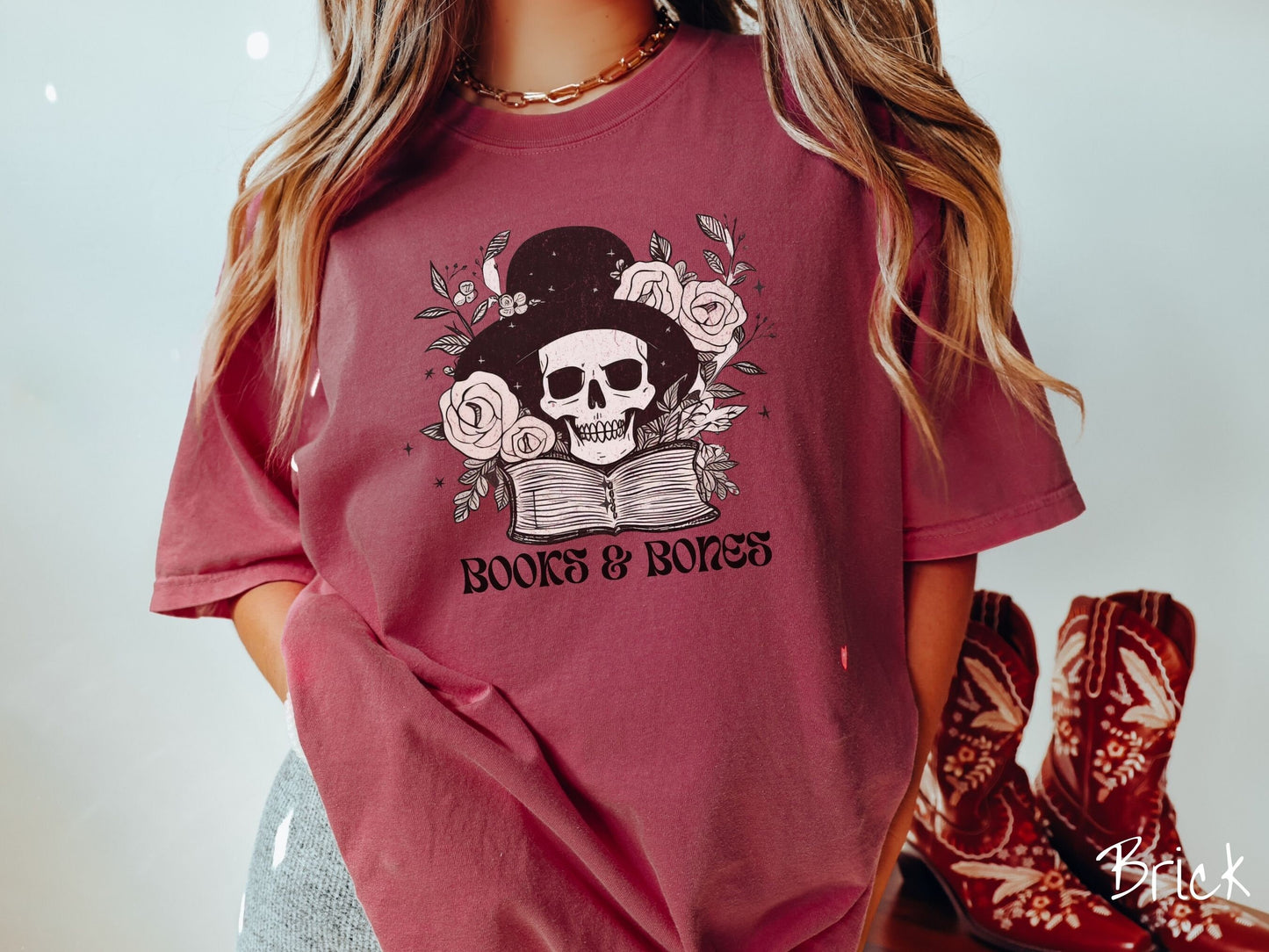 A woman wearing a cute, vintage brick colored Comfort Colors T-shirt with the text Books & Bones in black font. Above is a smiling skeleton wearing a black top hat surrounded by roses and leaves, and hovering above a thick, leather bound book.