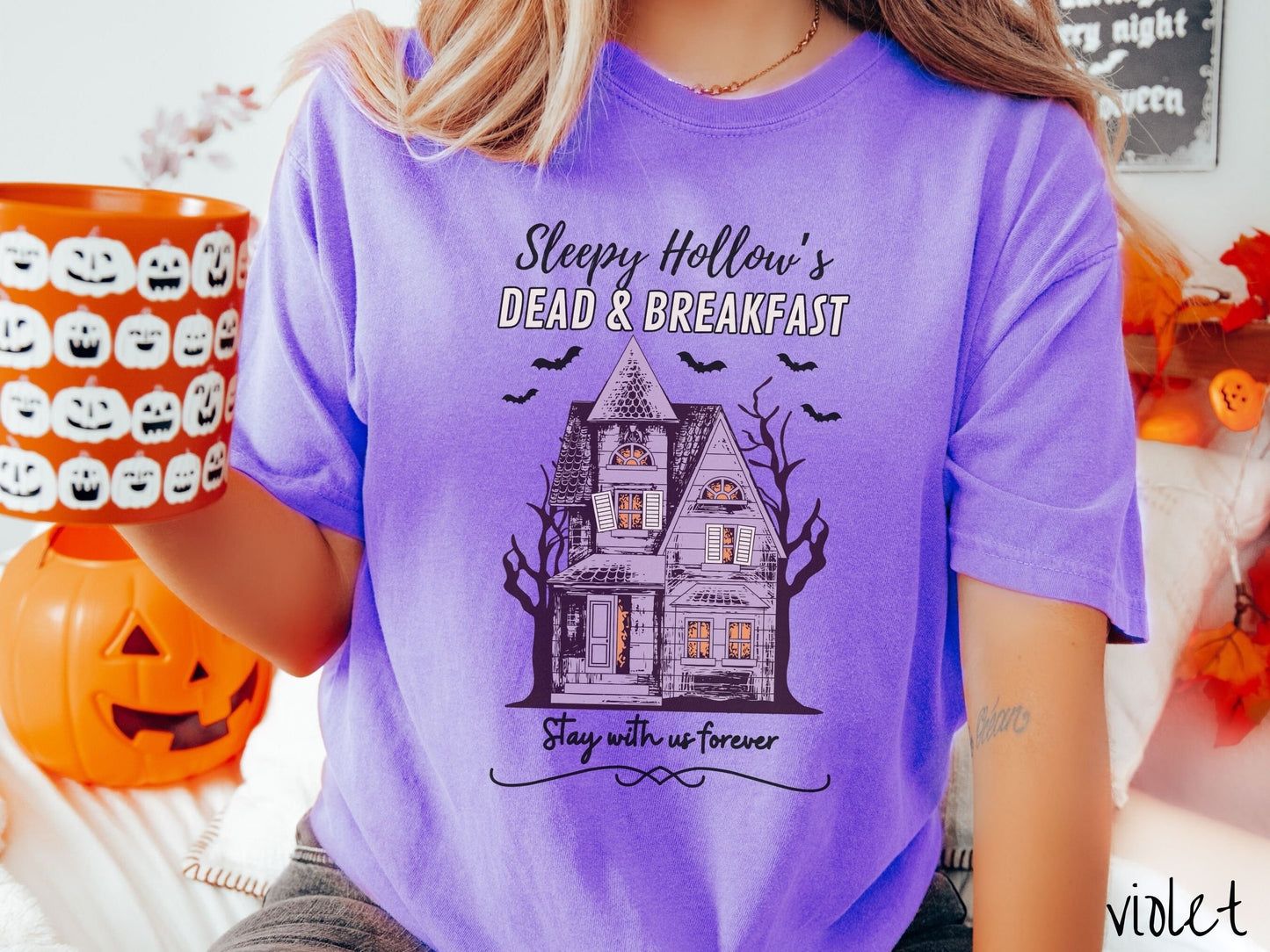 A woman wearing a cute, vintage violet colored Comfort Colors T-shirt with the text Sleepy Hollow’s Dead & Breakfast above then Stay with us forever below. In between the text is a spooky haunted house surrounded by black bats.