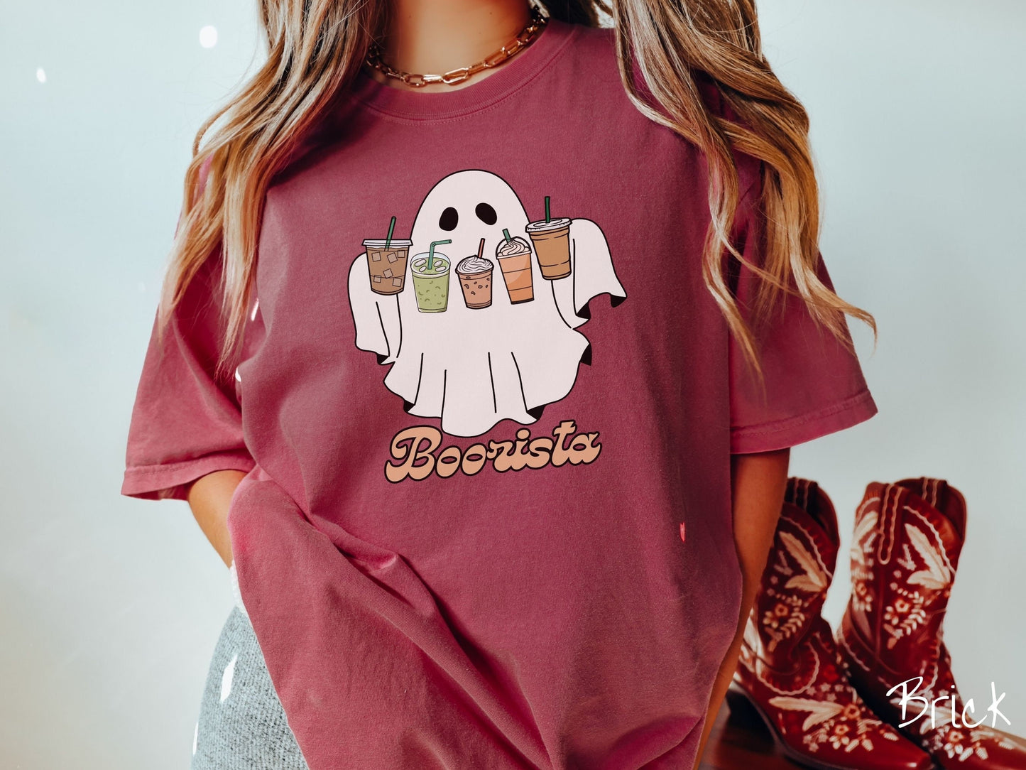A woman wearing a cute, vintage brick colored Comfort Colors T-shirt with the text Boorista along the bottom. Above is a white ghost holding an iced coffee, a green matcha tea, a frappuccino, a pumpkin spiced latte, and an iced mocha latte.