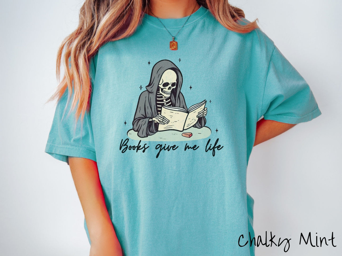 A woman wearing a cute, vintage chalky mint colored Comfort Colors T-shirt with the text Books give me life in black cursive font. Above is a smiling skeleton wearing a black hood reading a book surrounded by twinkling stars.