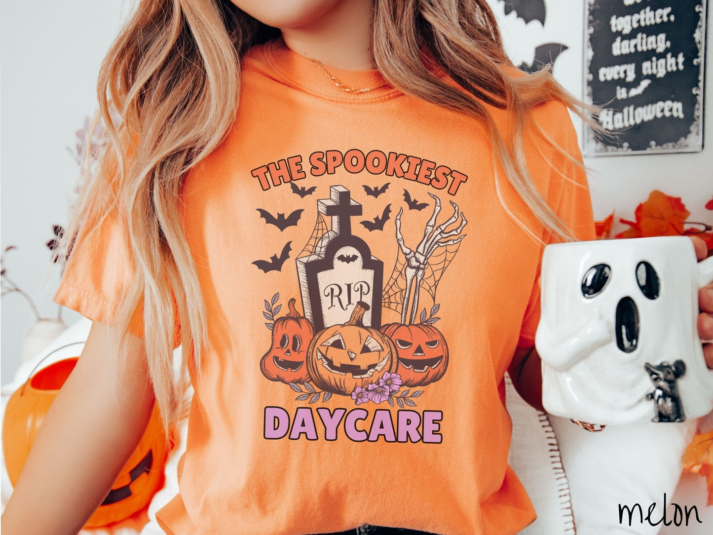 A woman wearing a cute, vintage melon colored Comfort Colors T-shirt with the text The Spookiest Daycare. There are three smiling carved pumpkins sitting in front of a tombstone with a skeleton arm coming out of the ground and black bats in the air.