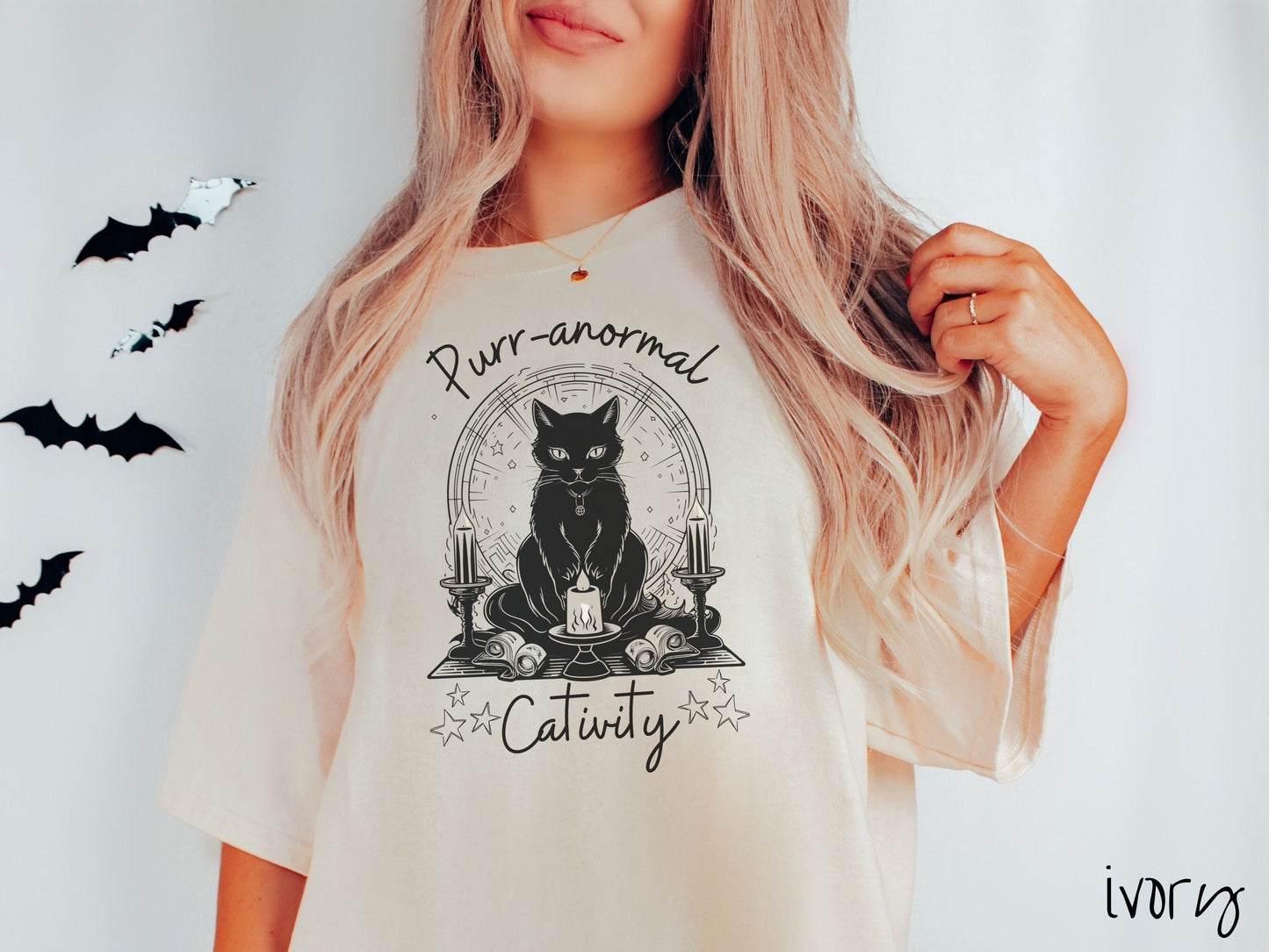 A woman wearing a cute, vintage ivory colored Comfort Colors T-shirt with text Purr-anormal Cativity in black font. There is a black cat sitting on an altar with candles and scrolls, staring straight forward with piercing eyes and wearing a necklace.