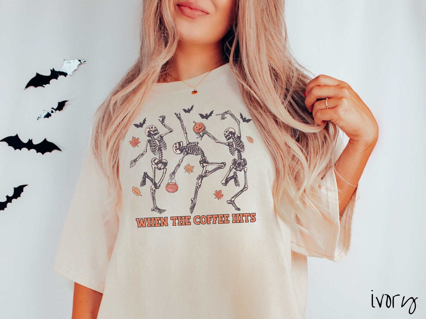 A woman wearing a cute, vintage ivory colored Comfort Colors T-shirt with the text When the Coffee Hits in orange font. Above are three dancing skeletons holding pumpkins and jackolantern trick or treat buckets with bats and leaves in the air.