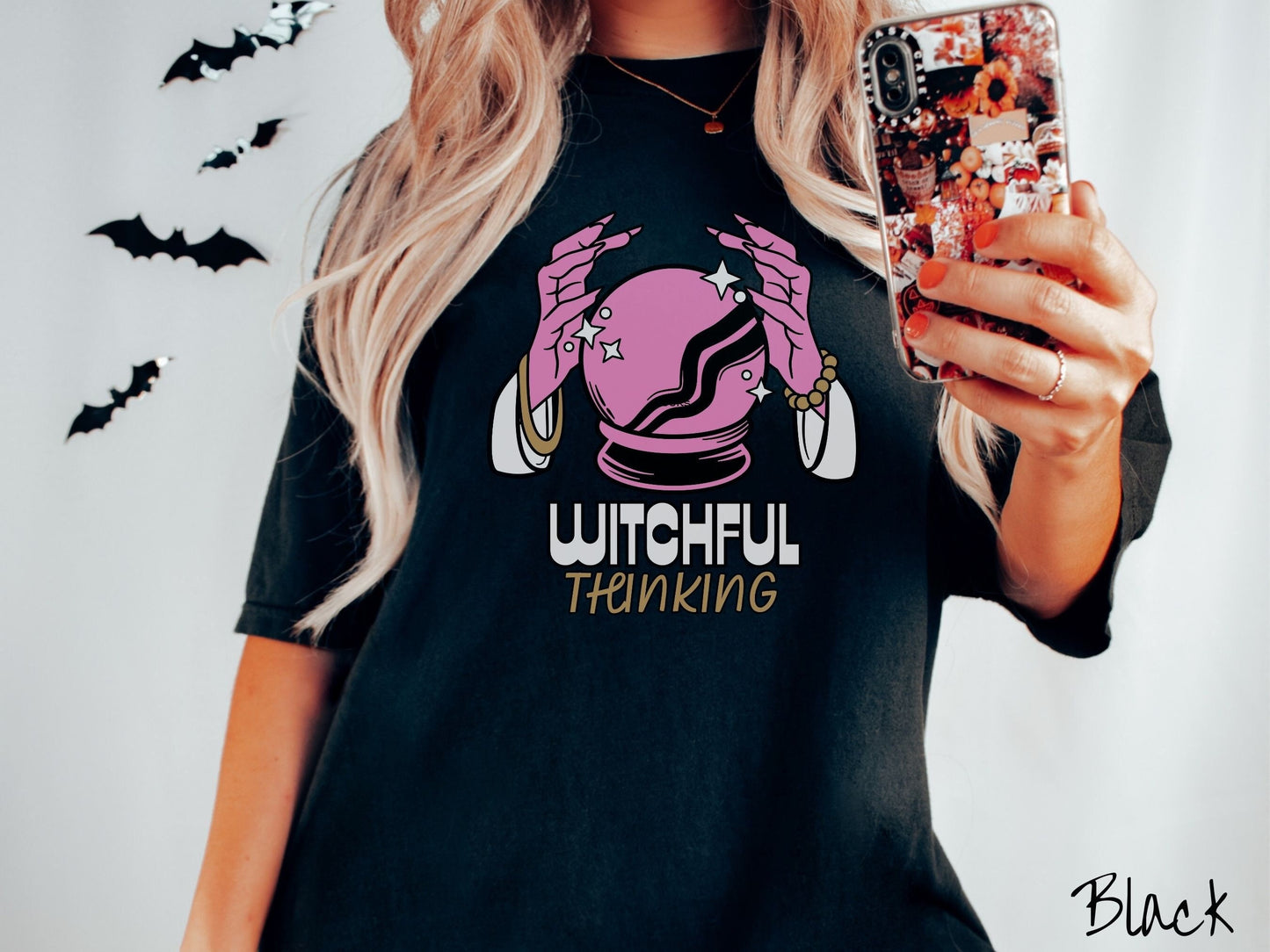 A woman wearing a cute, vintage black colored Comfort Colors T-shirt with the text Witchful Thinking in white and gold font. Above is a pink crystal ball, and there are two pink witch hands with jewelry hovering around the crystall ball.