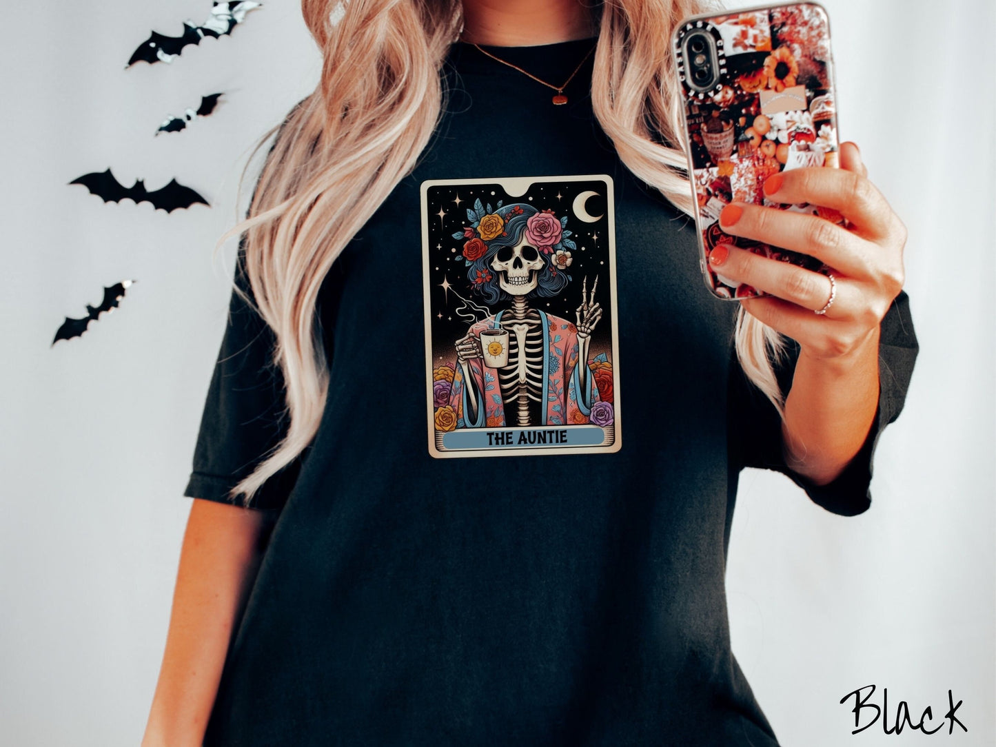 A woman wearing a cute, vintage black colored Comfort Colors T-shirt with text The Auntie along the bottom of a tarot card. Above is a picture of a female skeleton holding up a peace sign and wearing a flower robe holding a steaming cup of coffee.