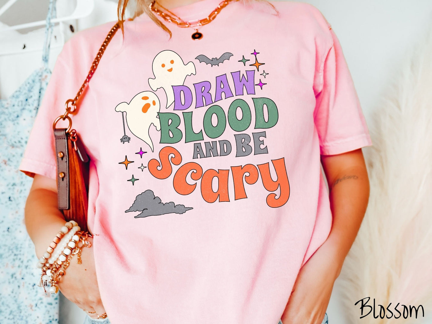 A woman wearing a cute, vintage blossom colored Comfort Colors T-shirt with the text Draw Blood and Be Scary in colorful font. There are orange-eyed white ghosts flying around and black bats and twinkling stars in the sky.