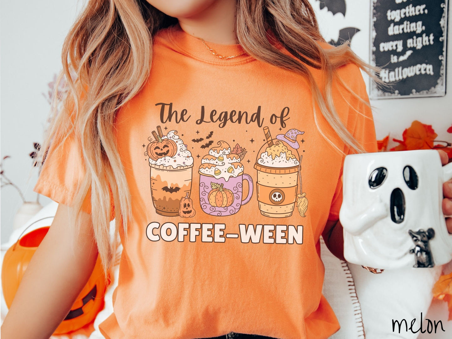 A woman wearing a cute, vintage melon colored Comfort Colors T-shirt with the text The legend of Coffee-ween. There are three iced coffees adorned with carved pumpkins, cinnamon sticks, whipped cream, bats, candy corn, whipped cream, and sprinkles.
