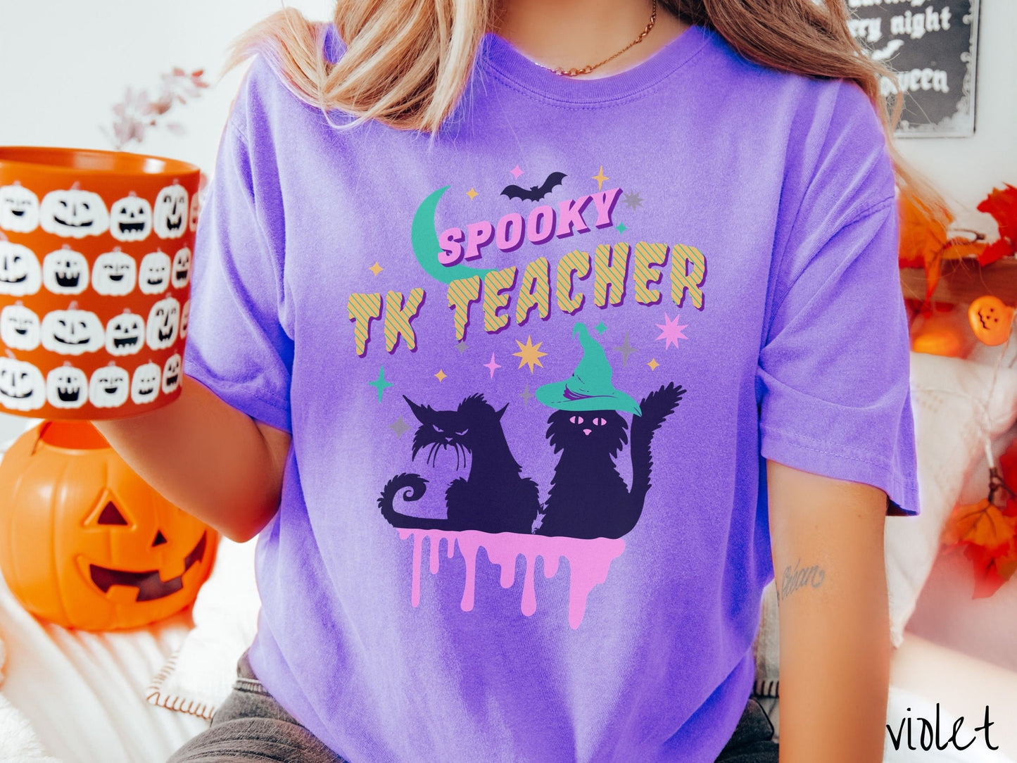 A woman wearing a cute, vintage violet colored Comfort Colors t-shirt with the text Spooky TK Teacher in pink and yellow font. Below are two black cats with pink eyes wearing witch hats sitting beneath colorful stars and a bright green moon.