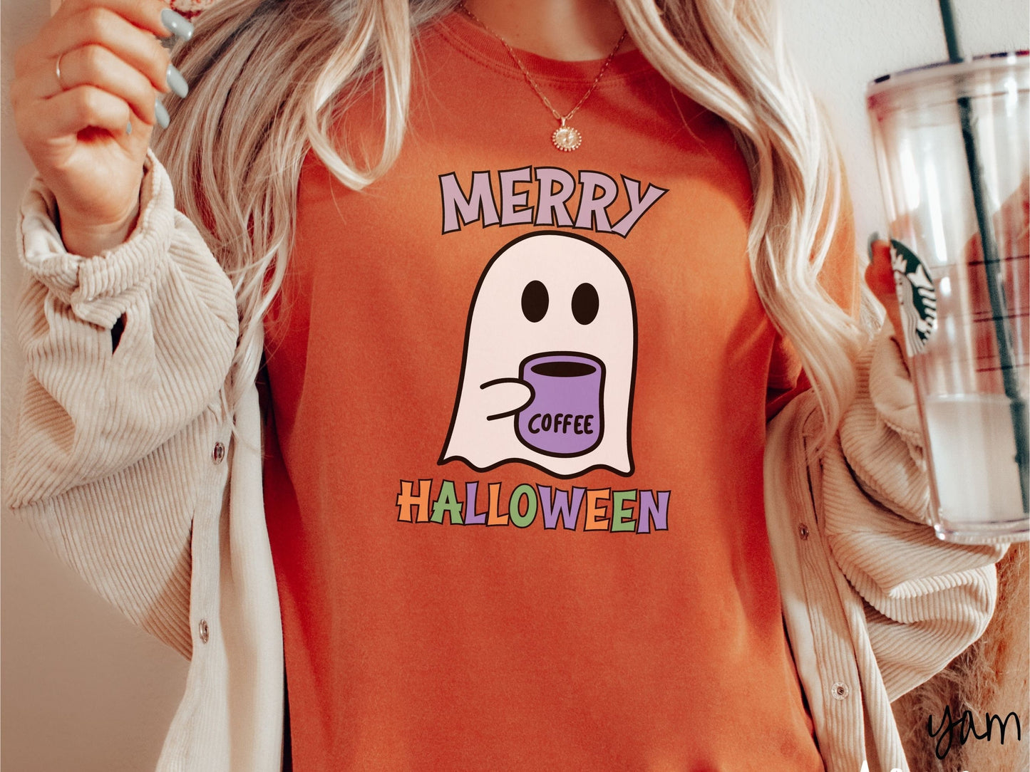 A woman wearing a cute, vintage yam colored Comfort Colors t-shirt with the text Merry Halloween in purple, orange, and green font. There is a cute ghost holding a coffee mug in between the text.