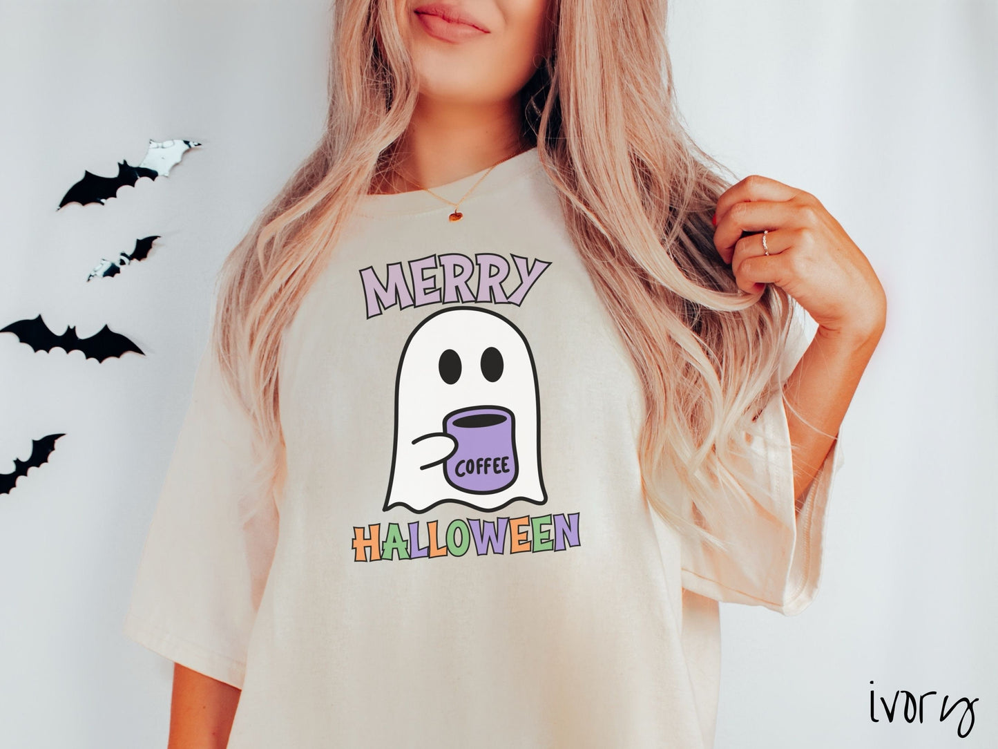 A woman wearing a cute, vintage ivory colored Comfort Colors t-shirt with the text Merry Halloween in purple, orange, and green font. There is a cute ghost holding a coffee mug in between the text.