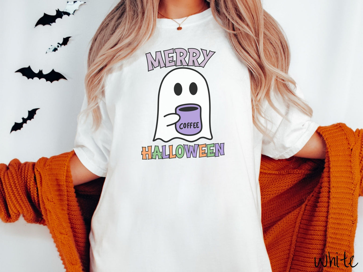 A woman wearing a cute, vintage white colored Comfort Colors t-shirt with the text Merry Halloween in purple, orange, and green font. There is a cute ghost holding a coffee mug in between the text.